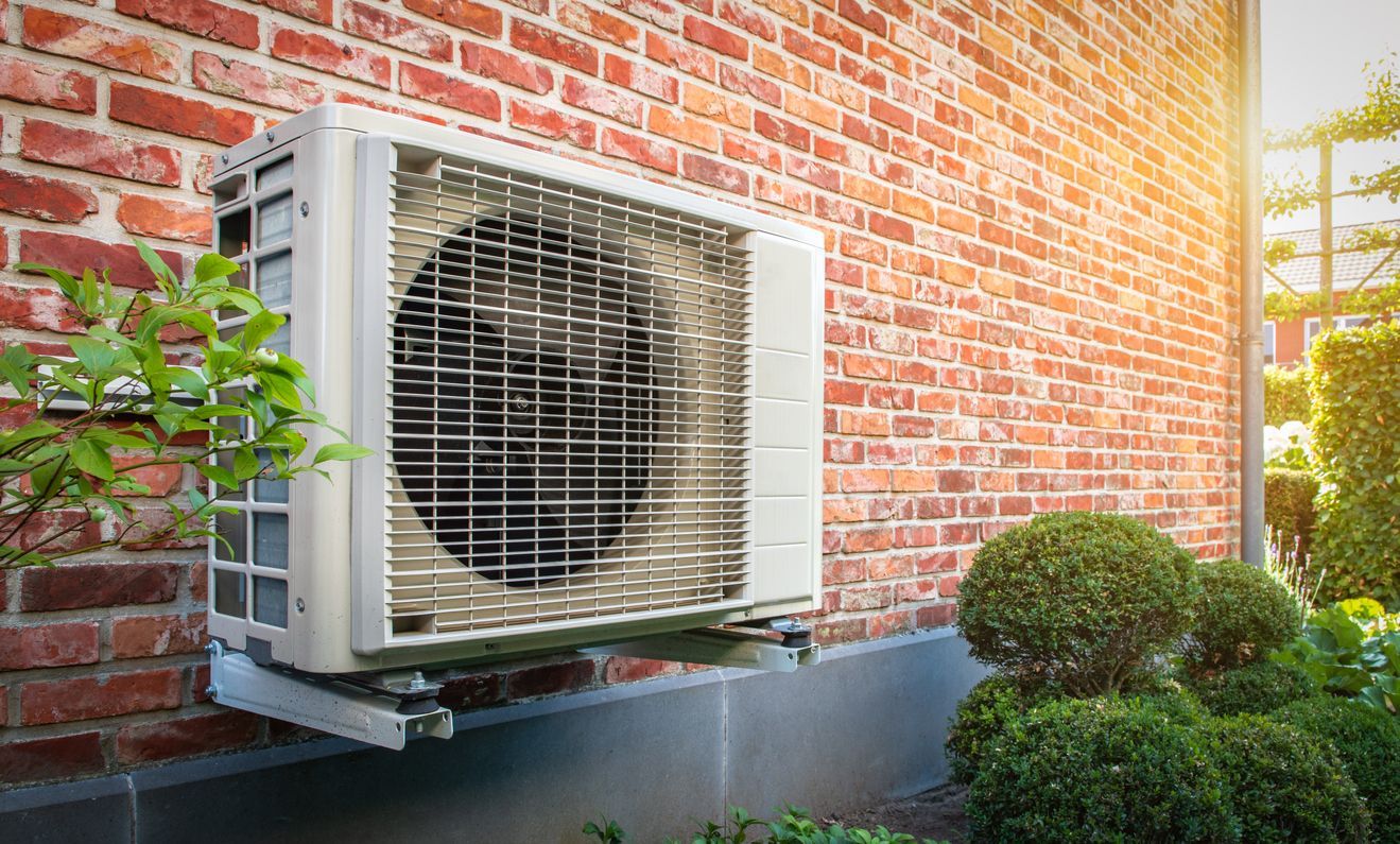 Air conditioning unit attached to a red brick wall, next to bushes.