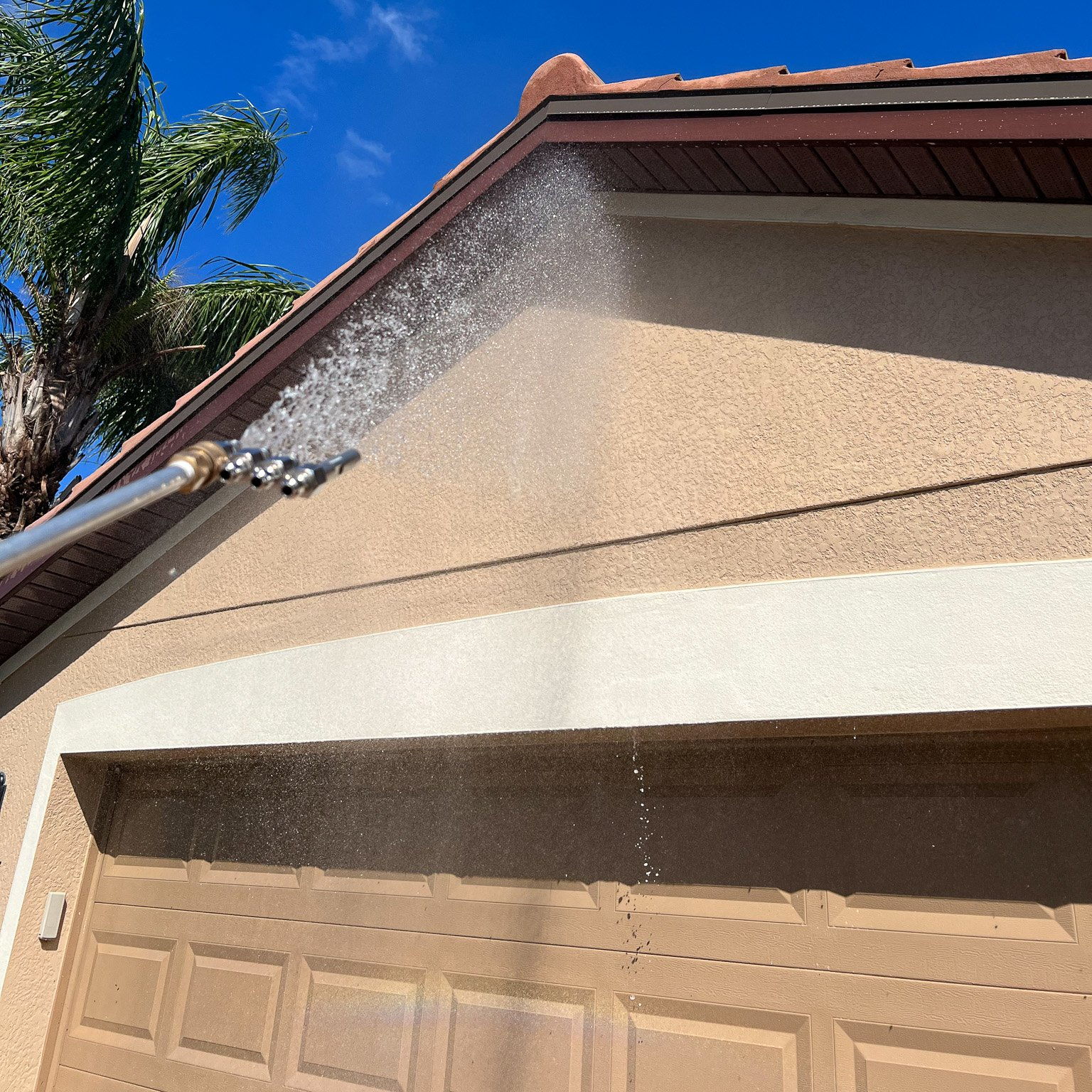 Person power washing the exterior of a tan house. Water spraying from a wand. Blue sky in the background.