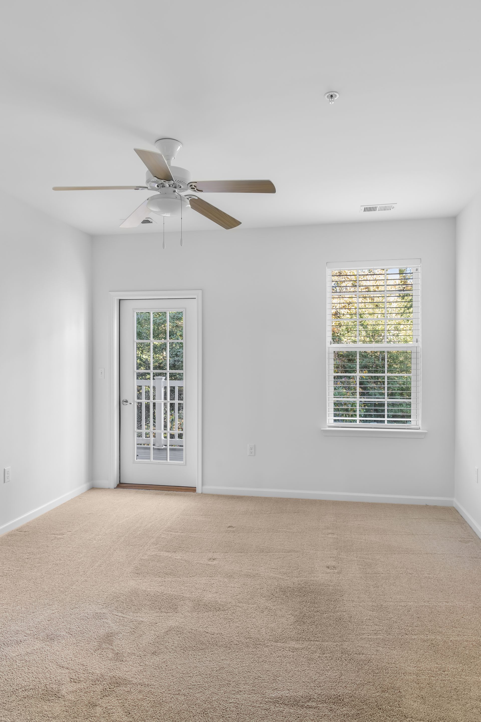 An empty room with white walls, beige carpet, a ceiling fan, a glass-paned door, and a window with blinds.