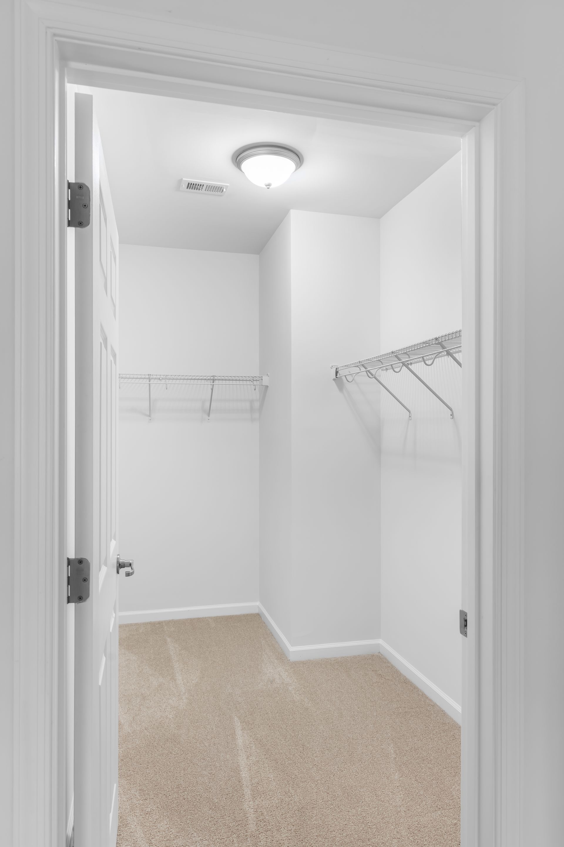 A walk-in closet with white walls, carpeted flooring, and mounted wire shelving on two sides under a ceiling light.