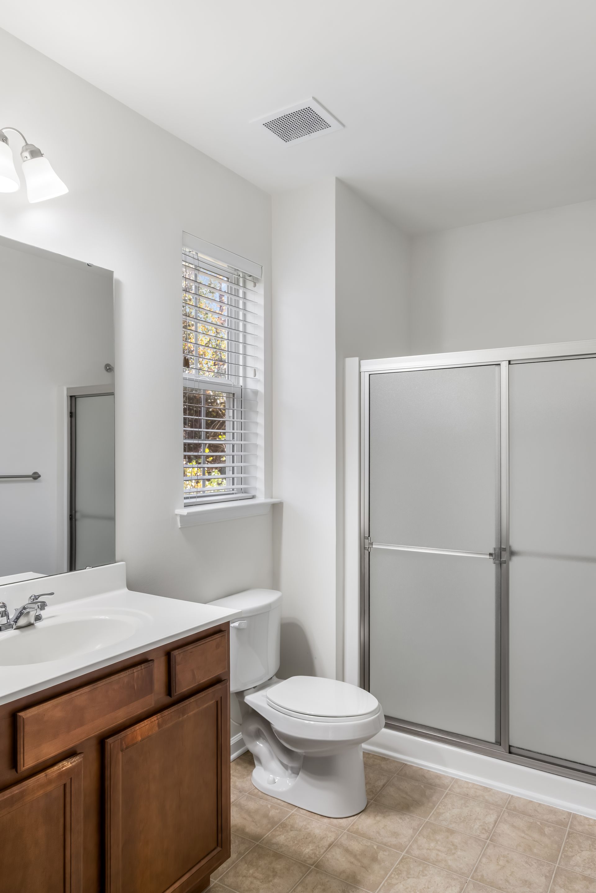 A neutral-toned bathroom with a wood vanity, white sink, toilet, frosted glass shower, and window with white blinds.