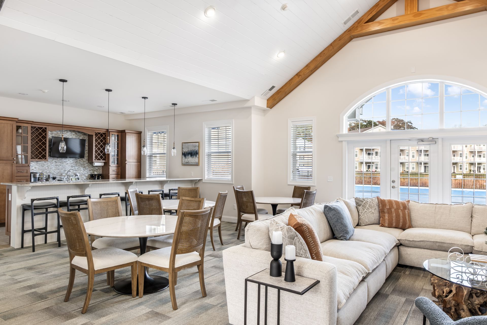A spacious, modern clubhouse lounge featuring white couches, dining sets, and a bar area with wooden cabinets.