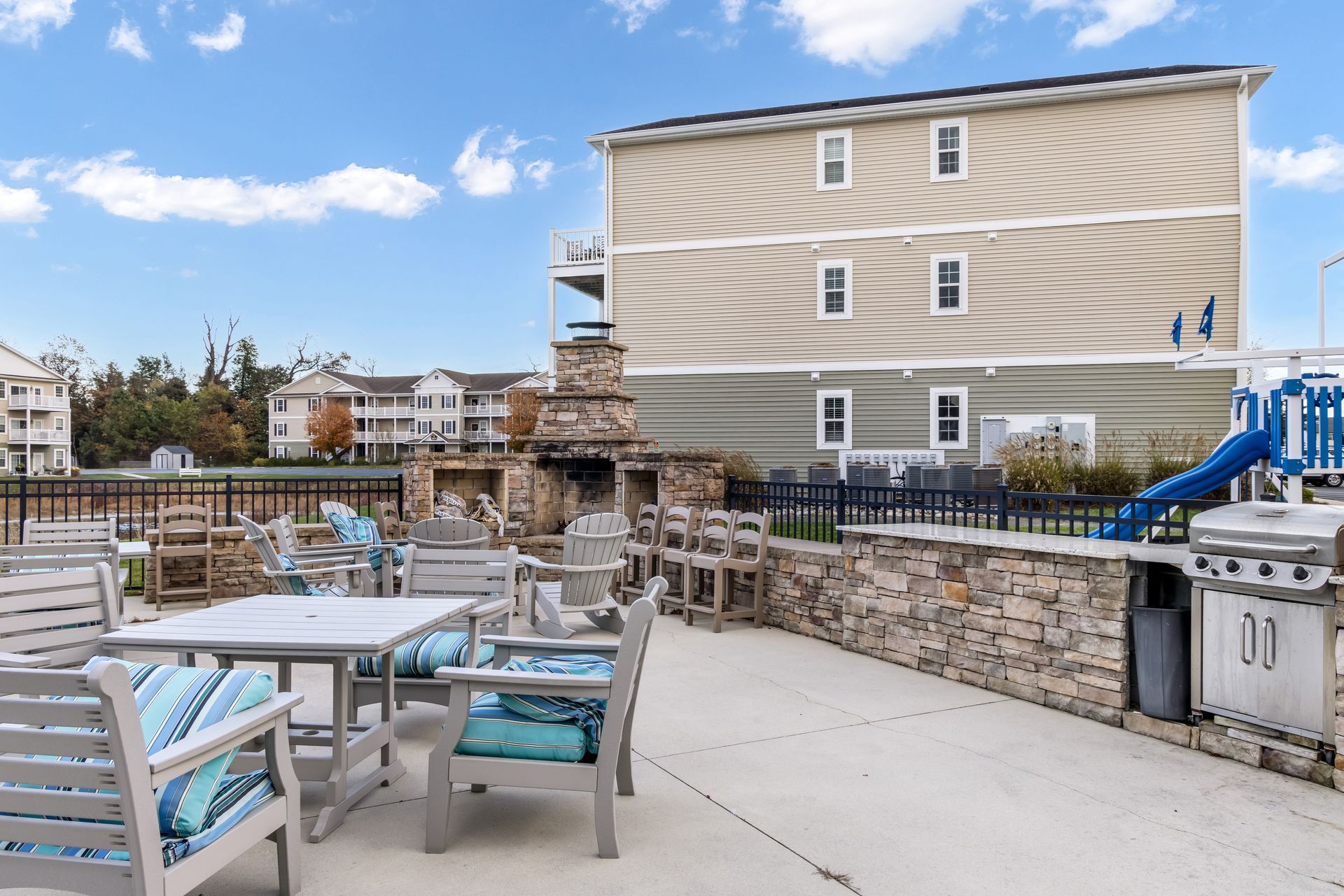Outdoor patio with stone fireplace, tables, chairs, and a grill near a large, light-colored residential building.