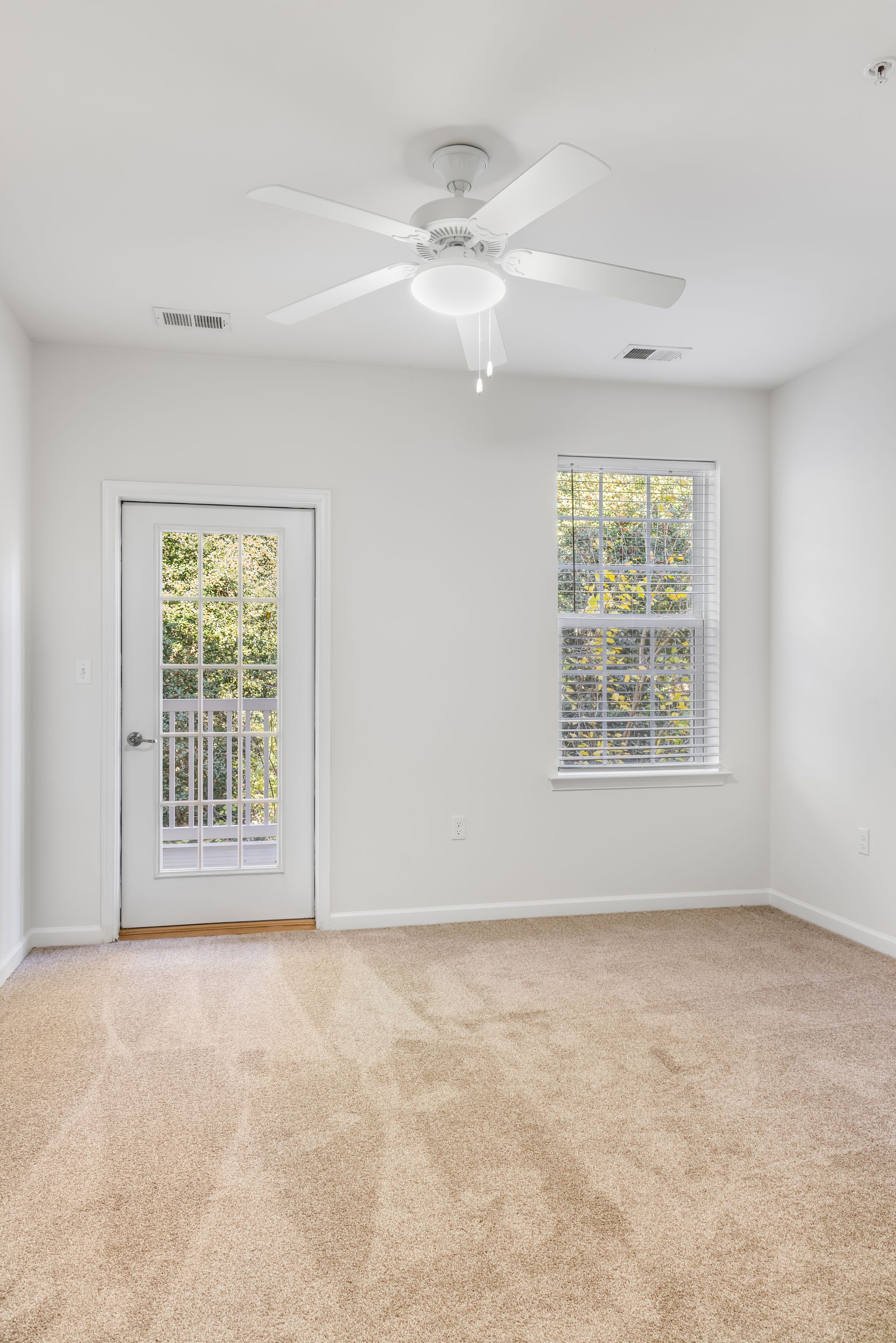 A bright, empty bedroom with white walls, beige carpet, a ceiling fan, a window, and a door leading outside.