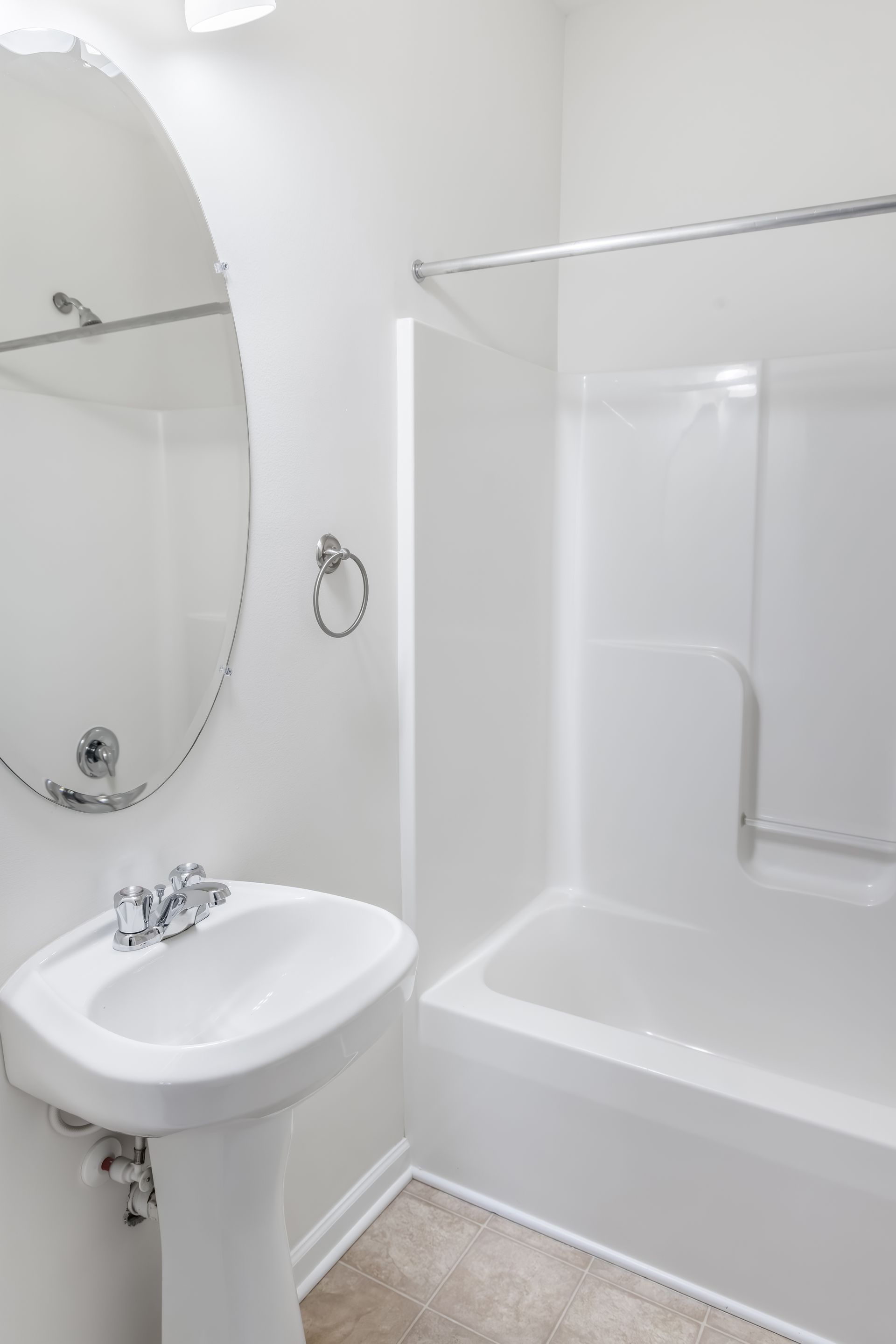 A bright, minimalist white bathroom with a pedestal sink, an oval wall mirror, and a shower-tub combination.