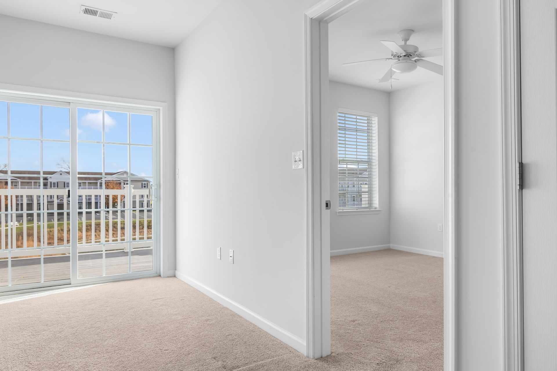 A bright, carpeted room with a sliding glass balcony door and an open doorway leading to a room with a ceiling fan.