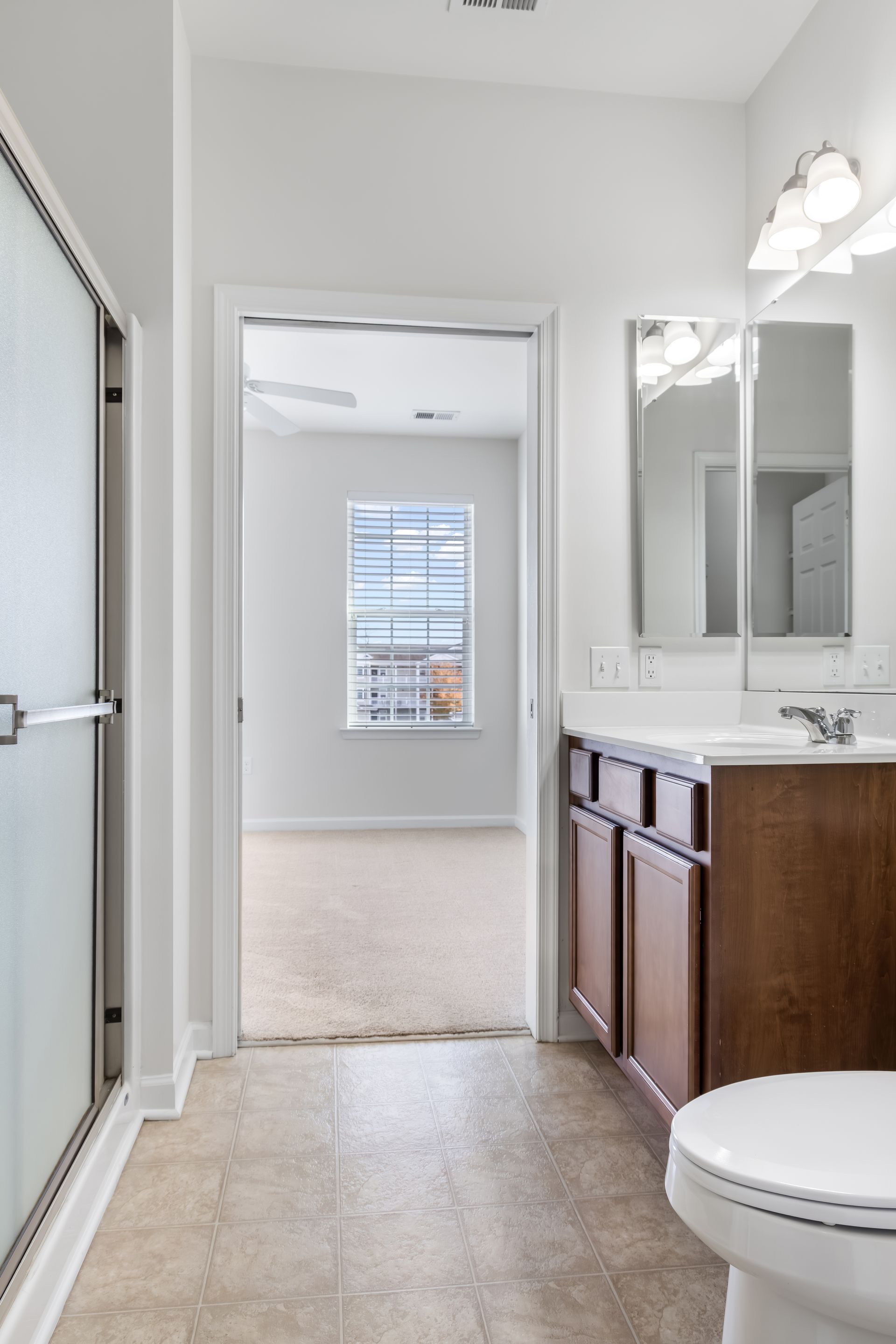Bathroom with a shower, vanity, and toilet, featuring a doorway leading to a carpeted bedroom with a window.