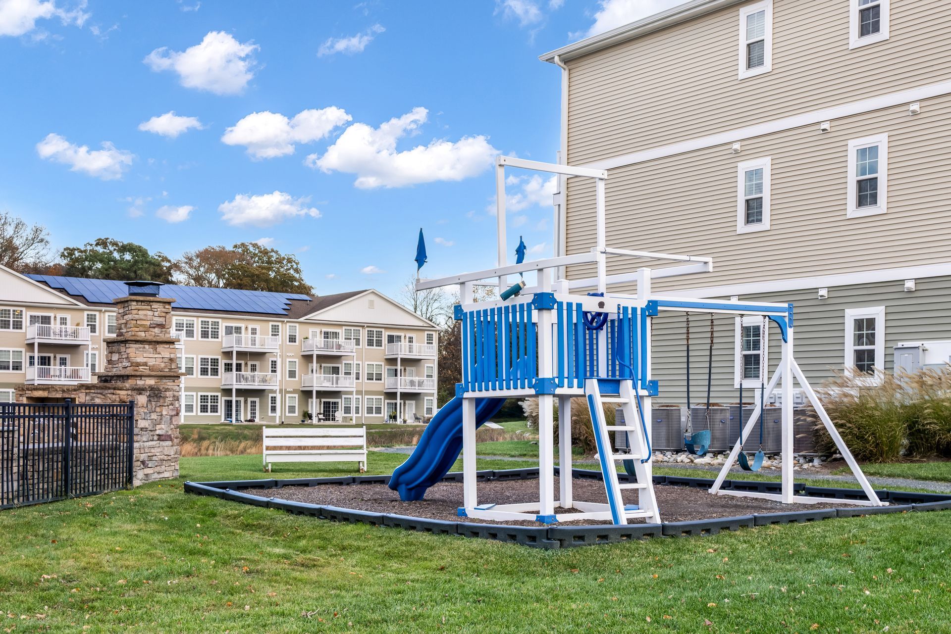 A blue and white playground set with a slide on a wood-chip ground, located in a courtyard between apartment buildings.