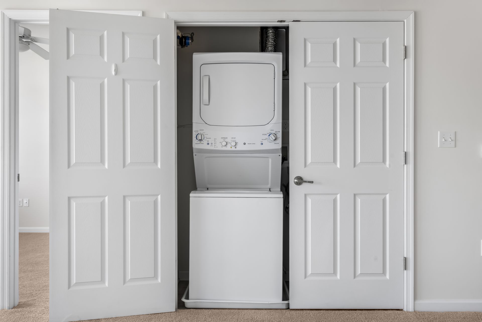 A white stacked washer and dryer unit inside an open closet, located in a room with neutral walls and carpeted floors.