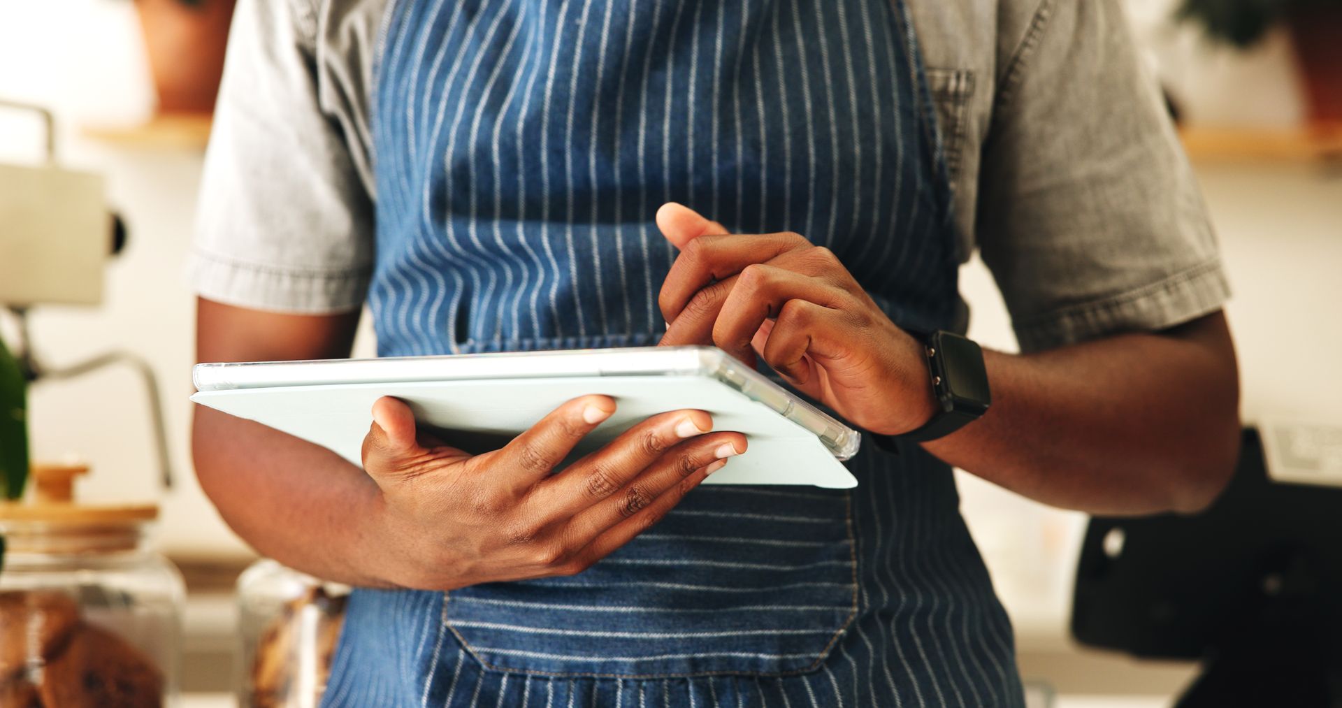 Person in blue apron using a tablet in a cafe setting, hands visible, looking at the screen.