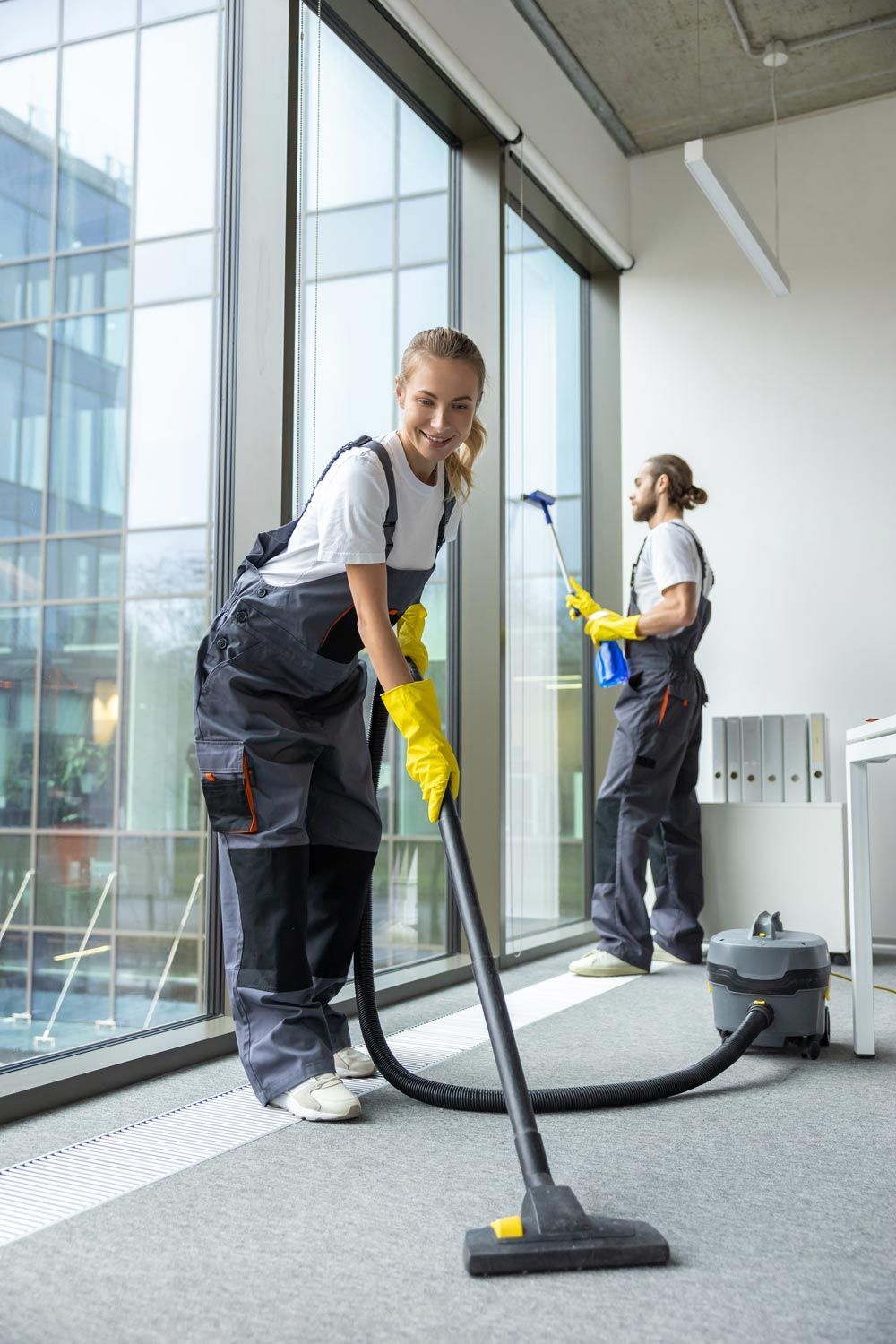 Two cleaners vacuuming a bright office with large windows and a carpeted floor