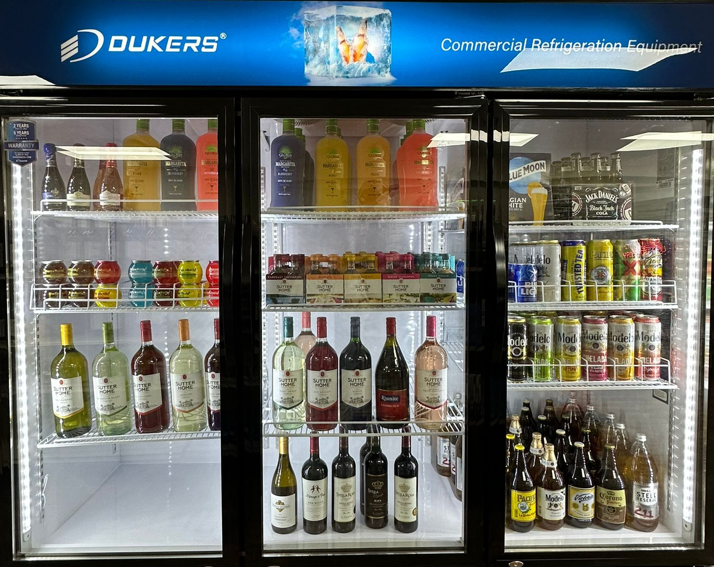 Three-door commercial refrigerator displaying various beverages: juices, wine, and beer.