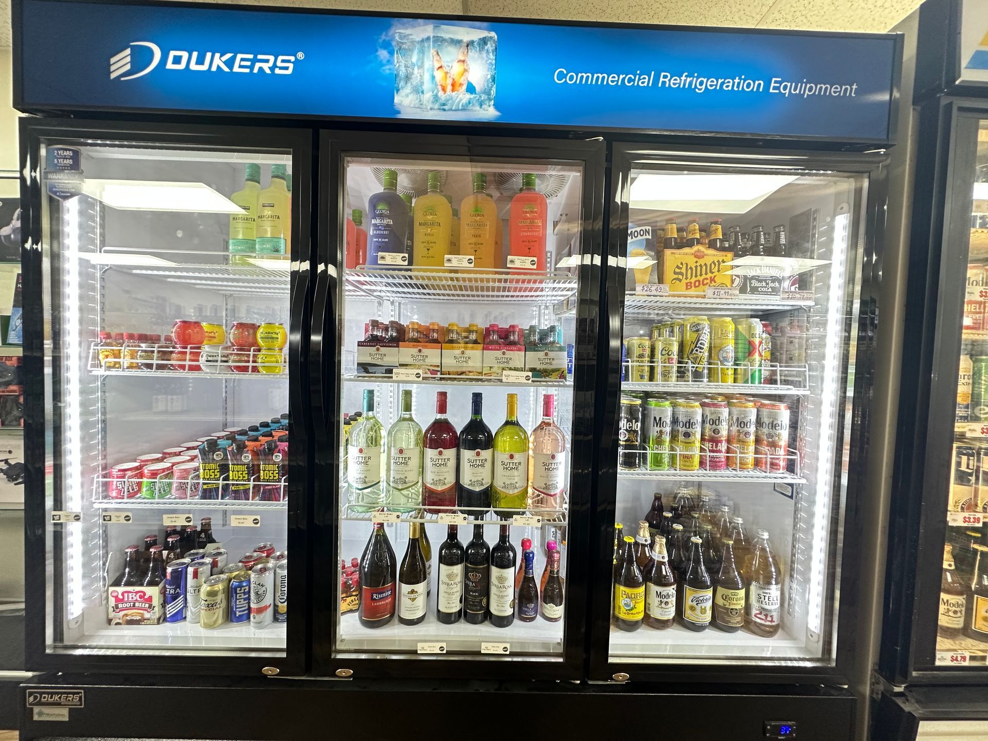 Refrigerated display case with beverages: soda, wine, beer. Commercial setting, lit interior.