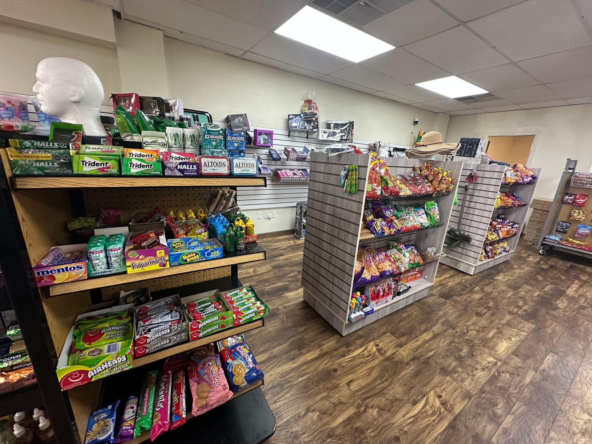Inside a convenience store, shelves filled with snacks and candy. Wooden and white shelving, wood-look floor.