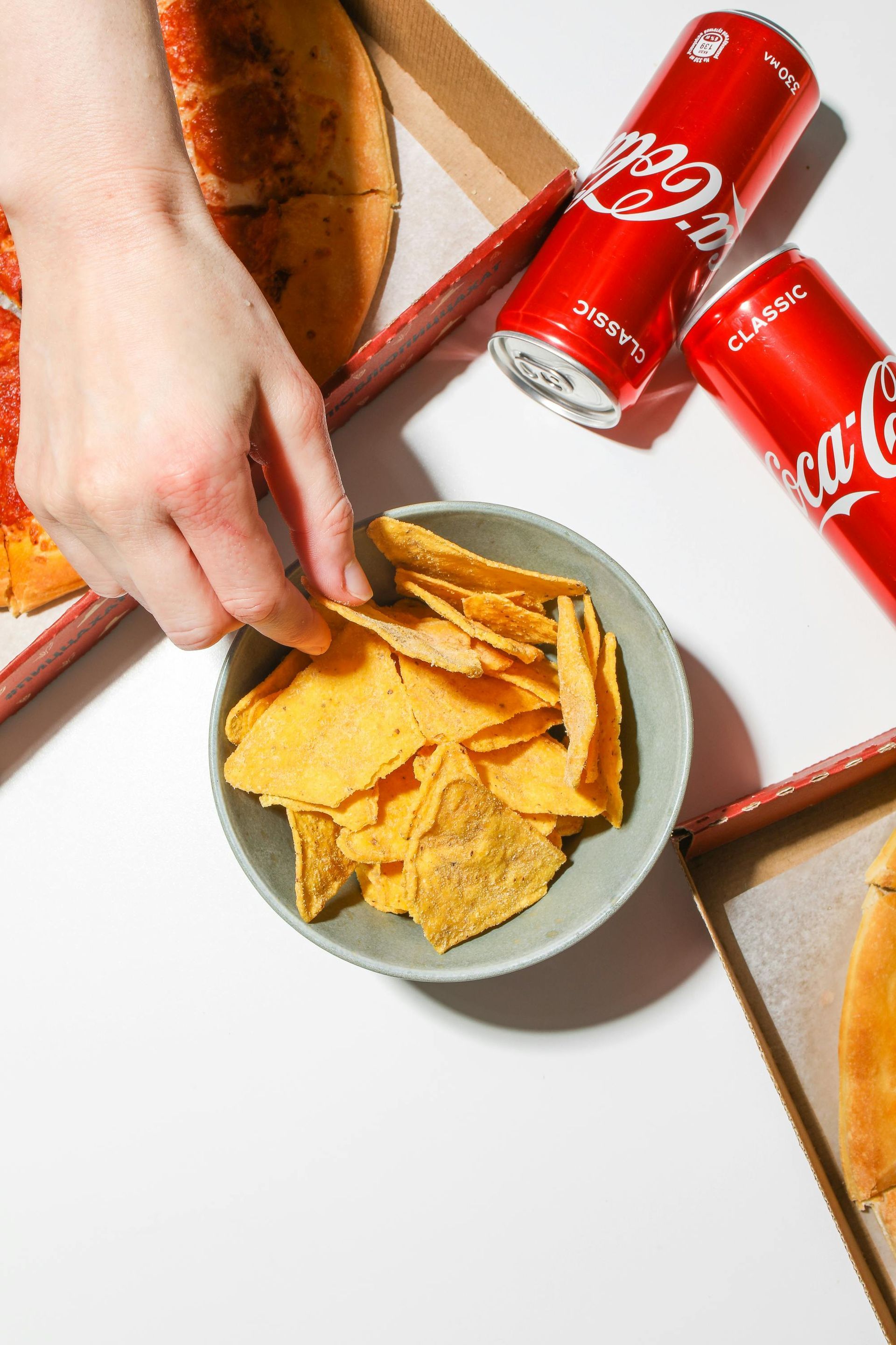 Hand reaching for a tortilla chip from a bowl, pizza and Coca-Cola cans are nearby.