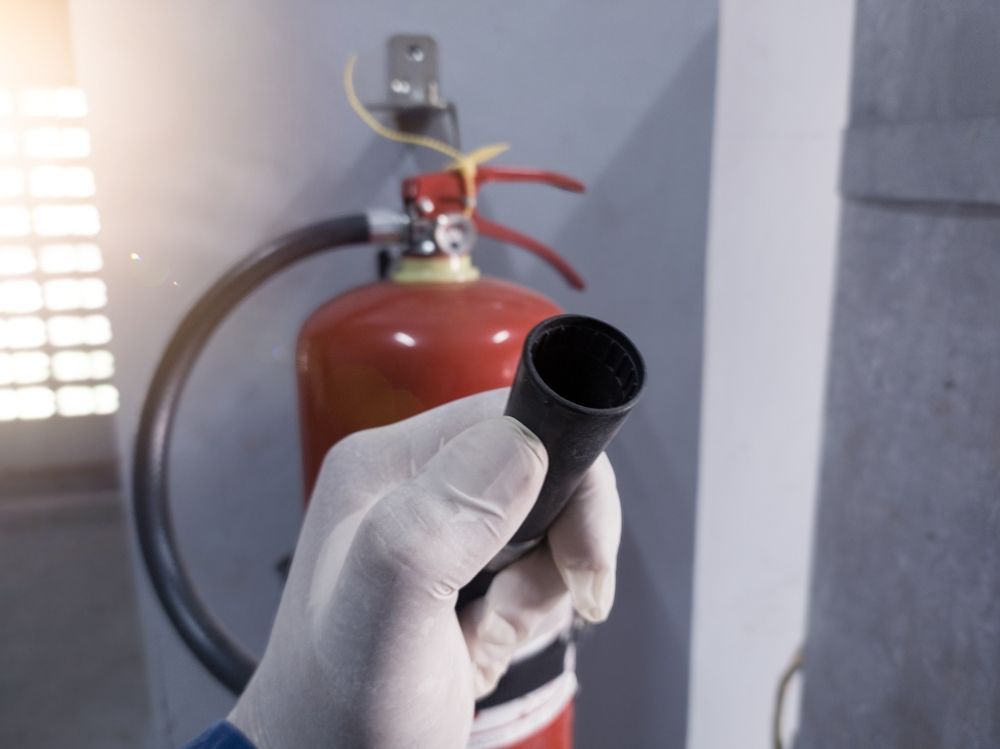 Gloved hand holding a black nozzle near a red fire extinguisher mounted on a wall