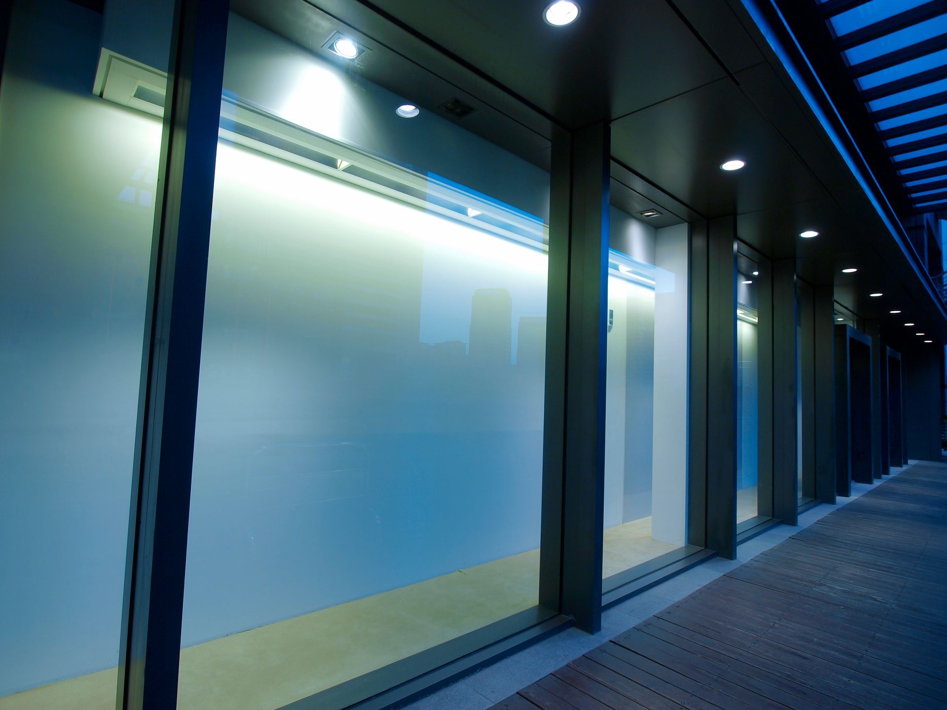Modern building exterior with glass windows, overhead lighting, and a walkway. Blue and gray tones.