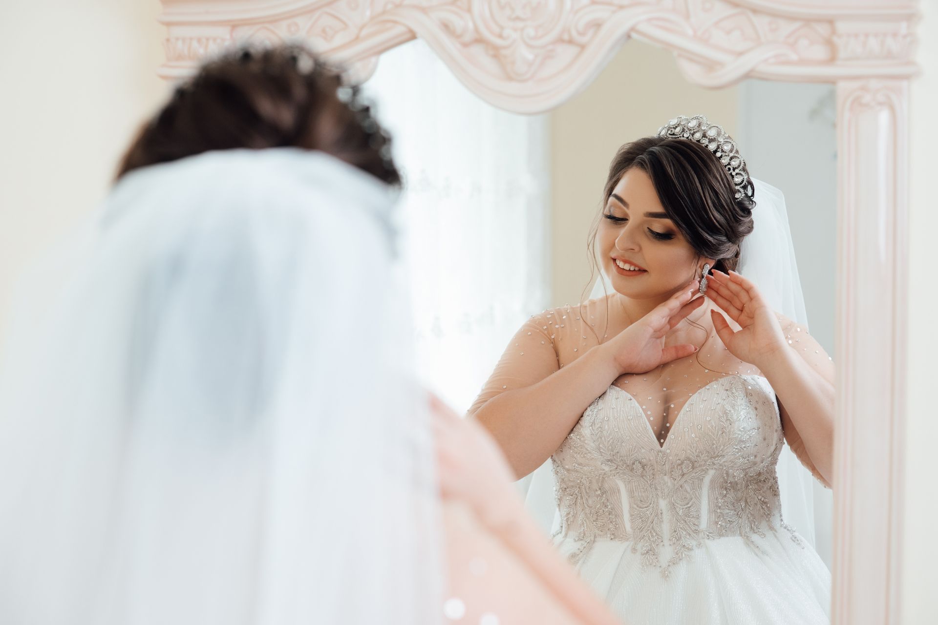 Bride putting on earrings in a mirror; she wears a tiara, veil, and beaded wedding dress.