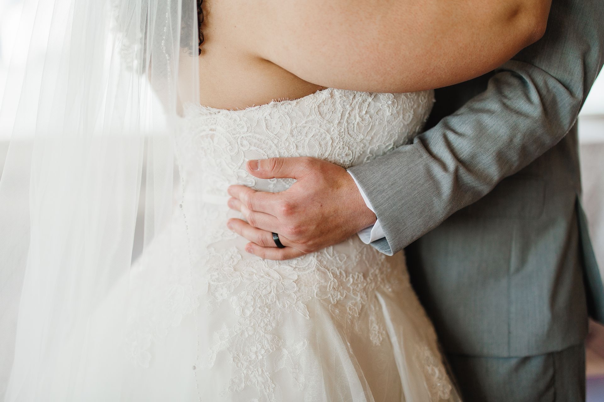 Bride in white lace dress embraced by person in gray suit.