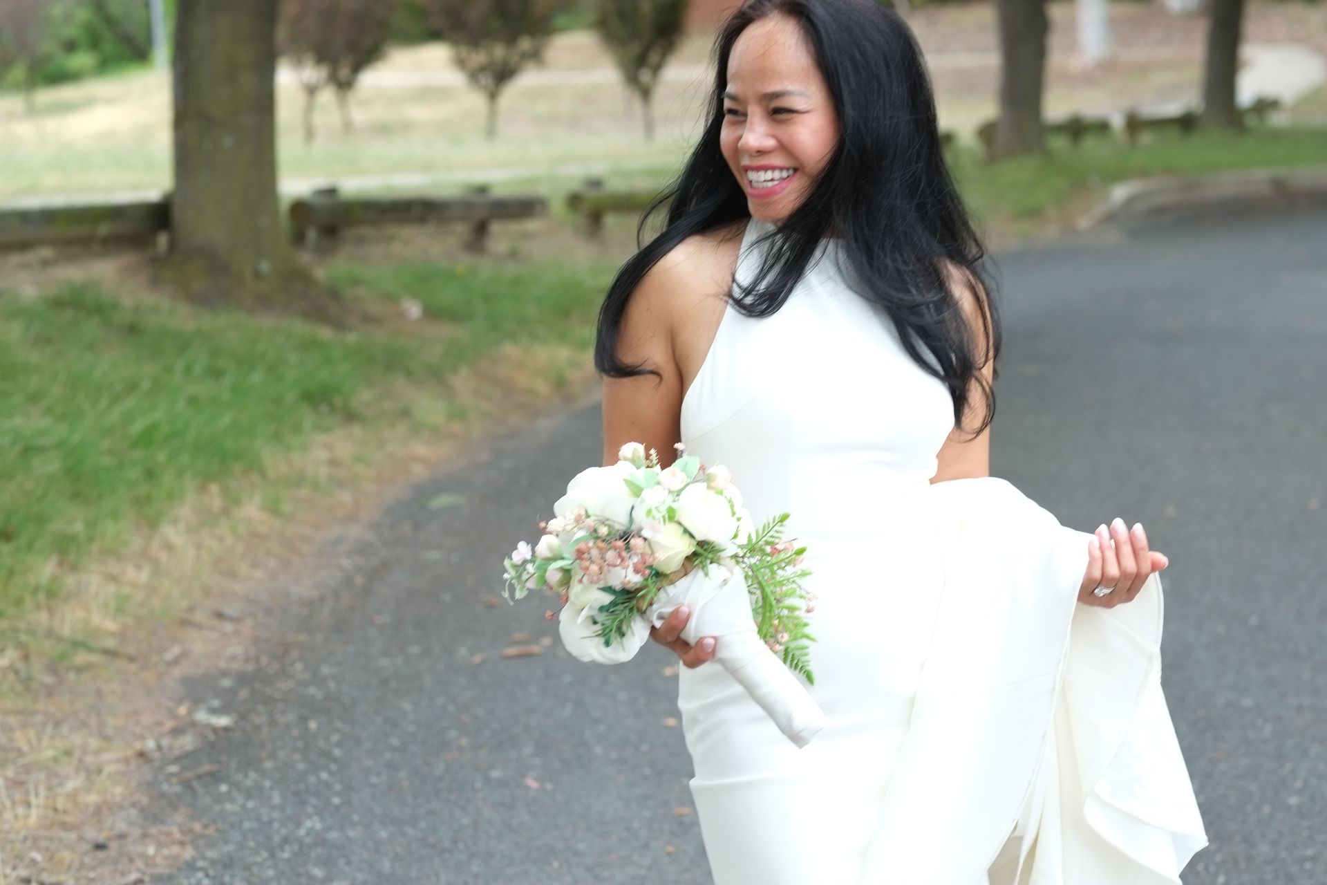 Woman in white dress smiles, holds bouquet and jacket, walks on path outdoors.