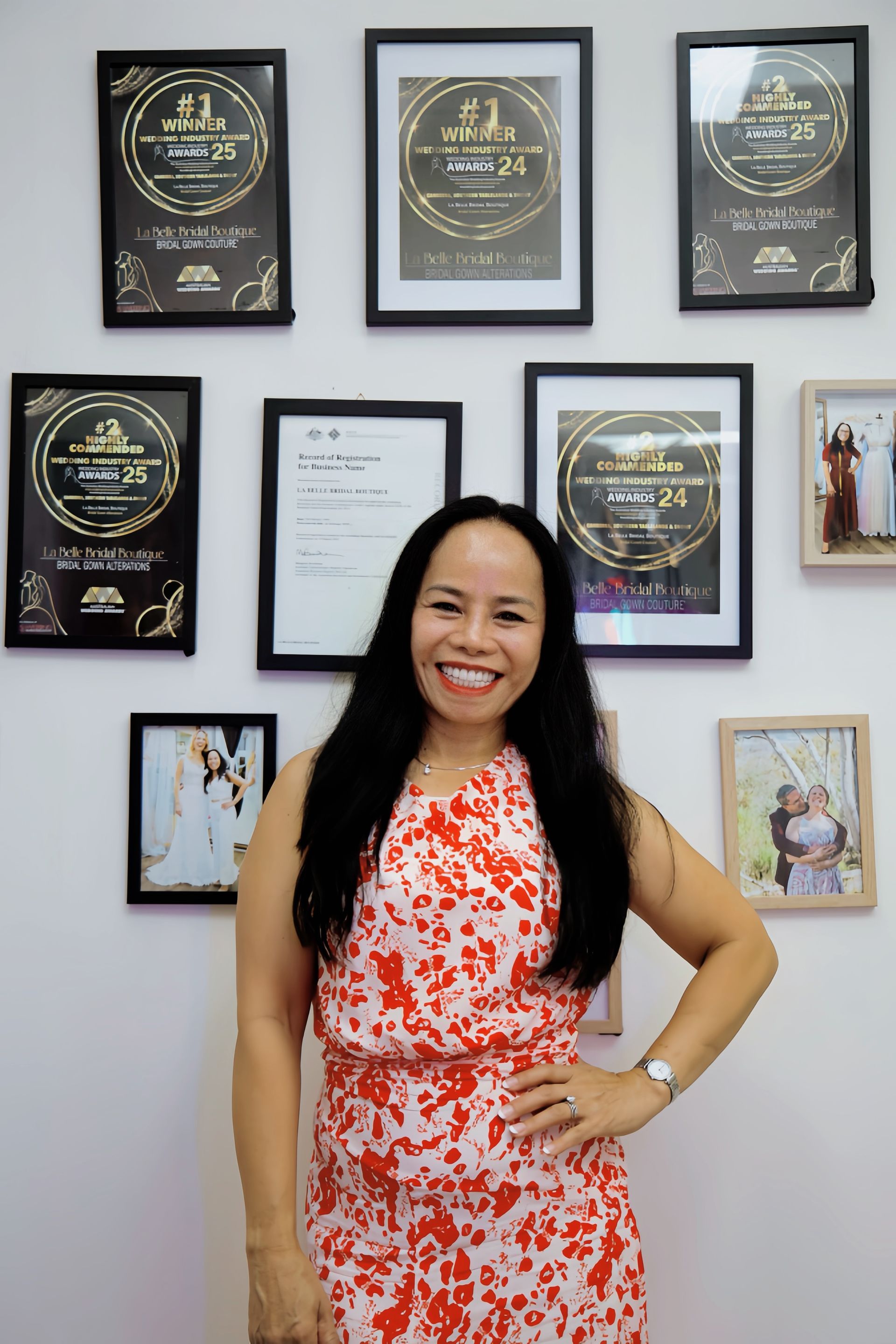 Woman in red and white dress smiles, posing by framed awards on a white wall.