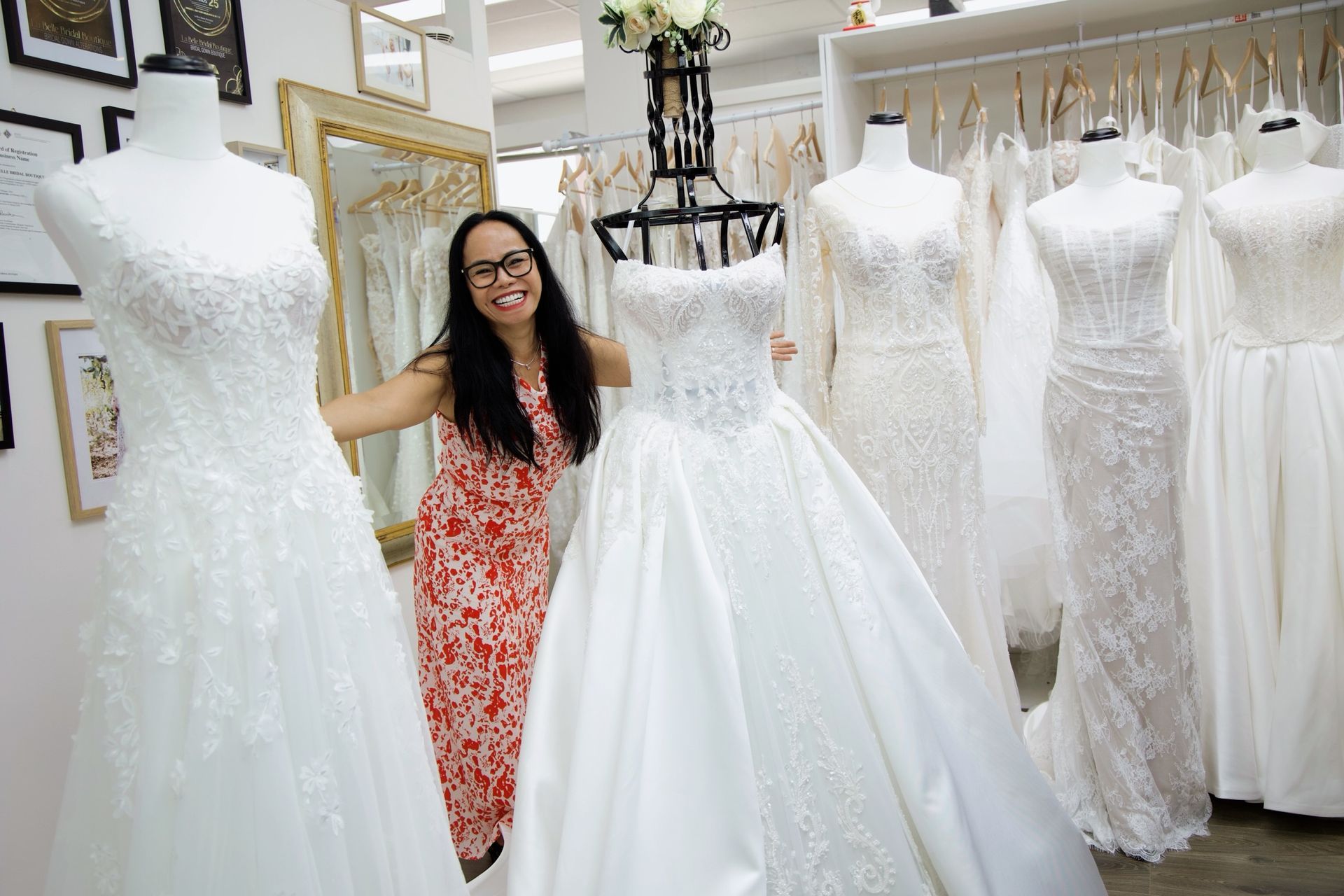 Woman in dress shop, surrounded by wedding dresses on mannequins. Smiling, she touches a satin gown.