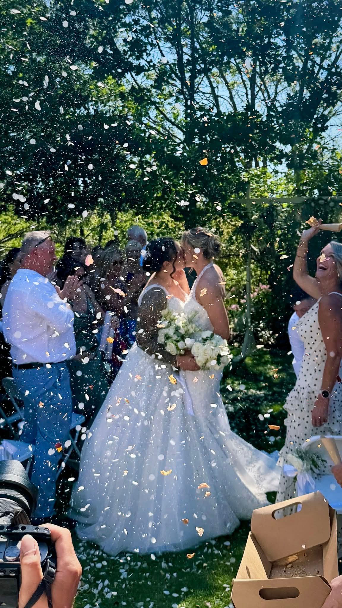 Two brides embrace under confetti, surrounded by guests in a garden setting.