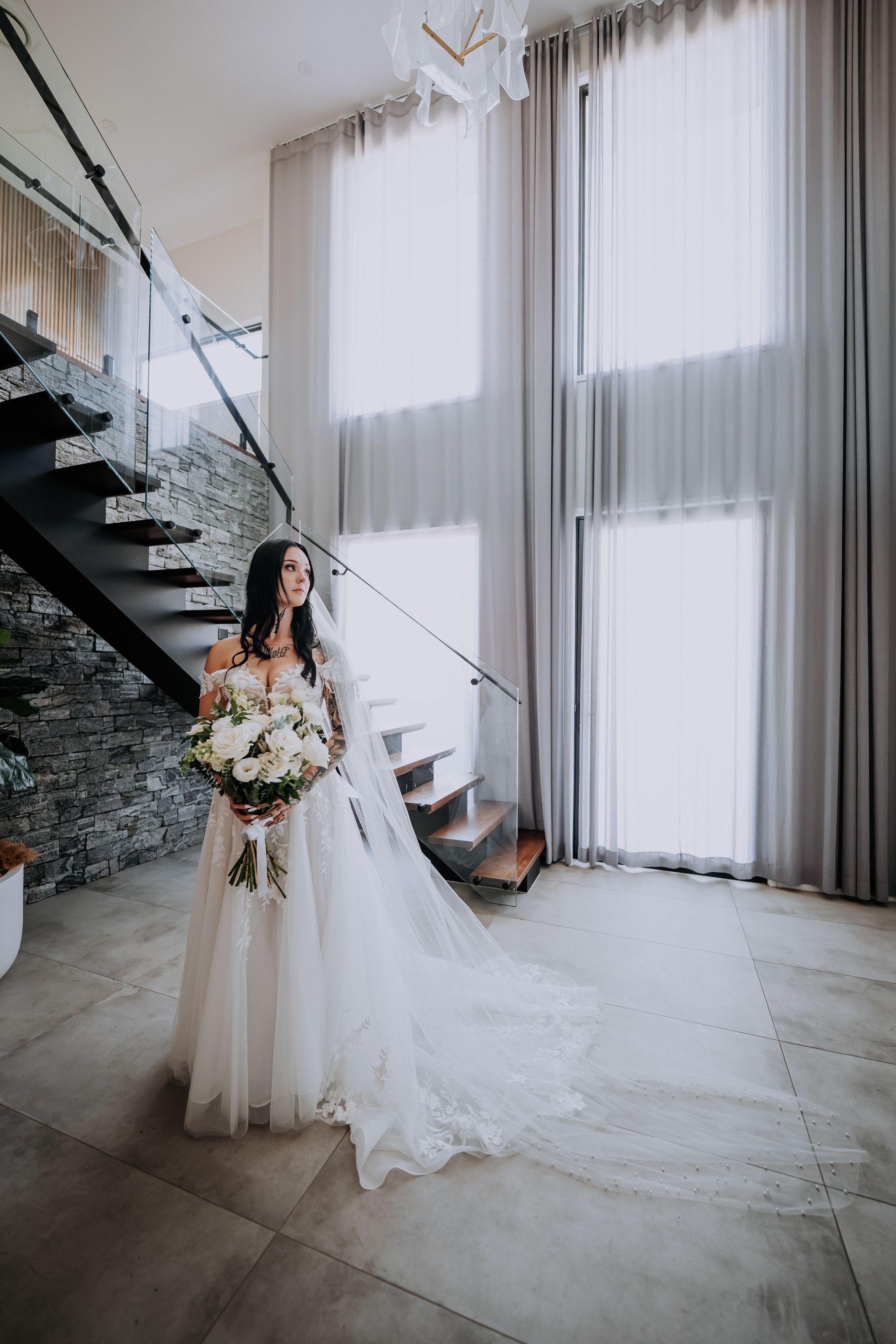 Bride in white gown, holding flowers, stands near staircase and window with sheer curtains.