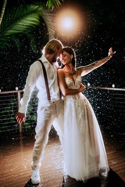 Couple dancing in the rain on a wooden deck at night; woman in a white wedding dress, man in white shirt and pants.