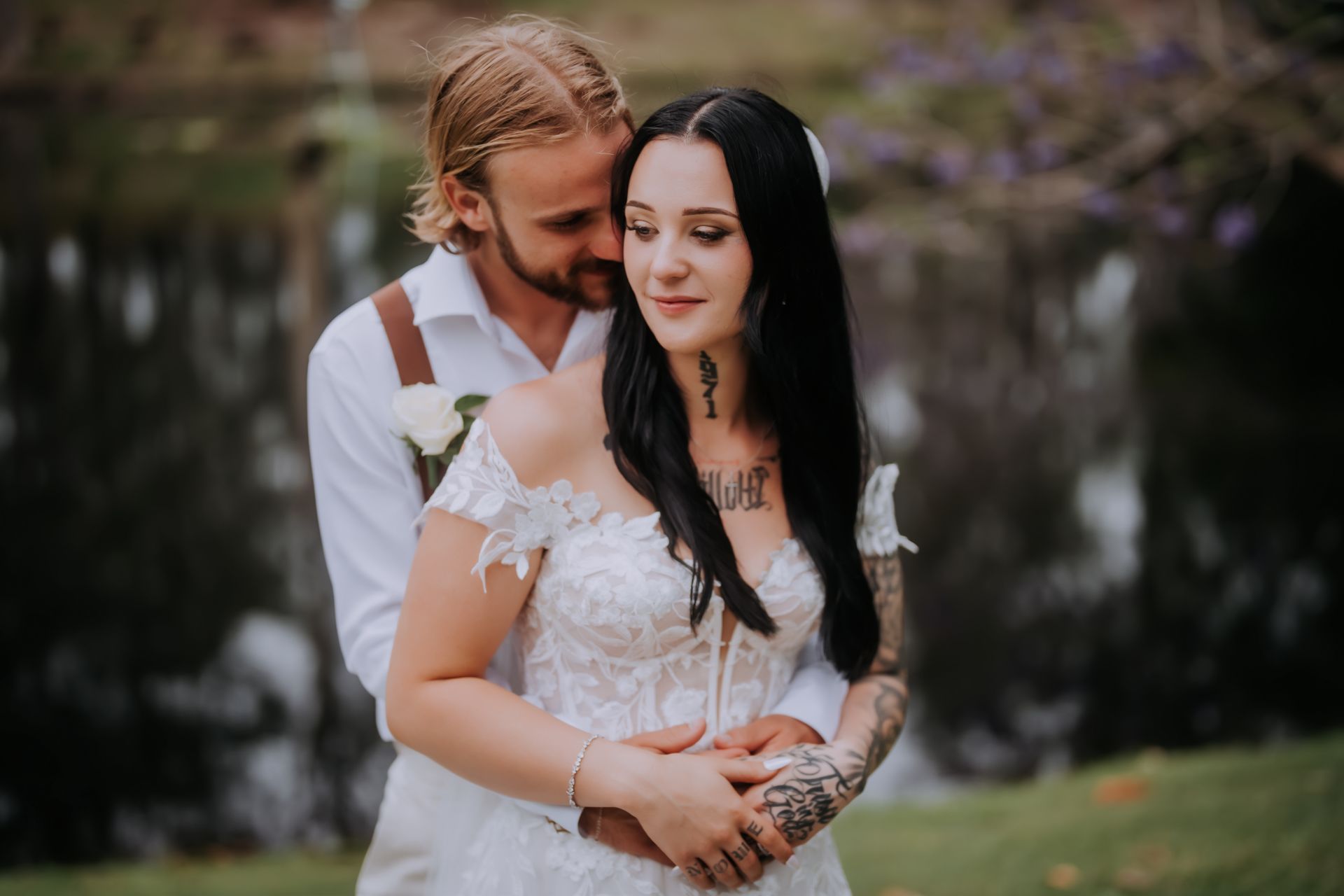 Newlyweds embrace outdoors: The bride in white dress, tattoos, and the groom with blonde braids.
