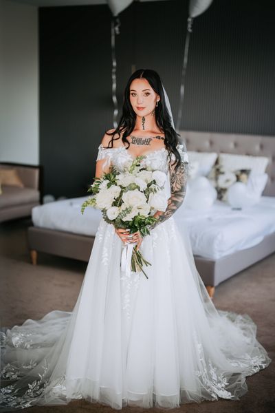 Bride in a white wedding gown, holding flowers. She has arm tattoos, standing in a bedroom.