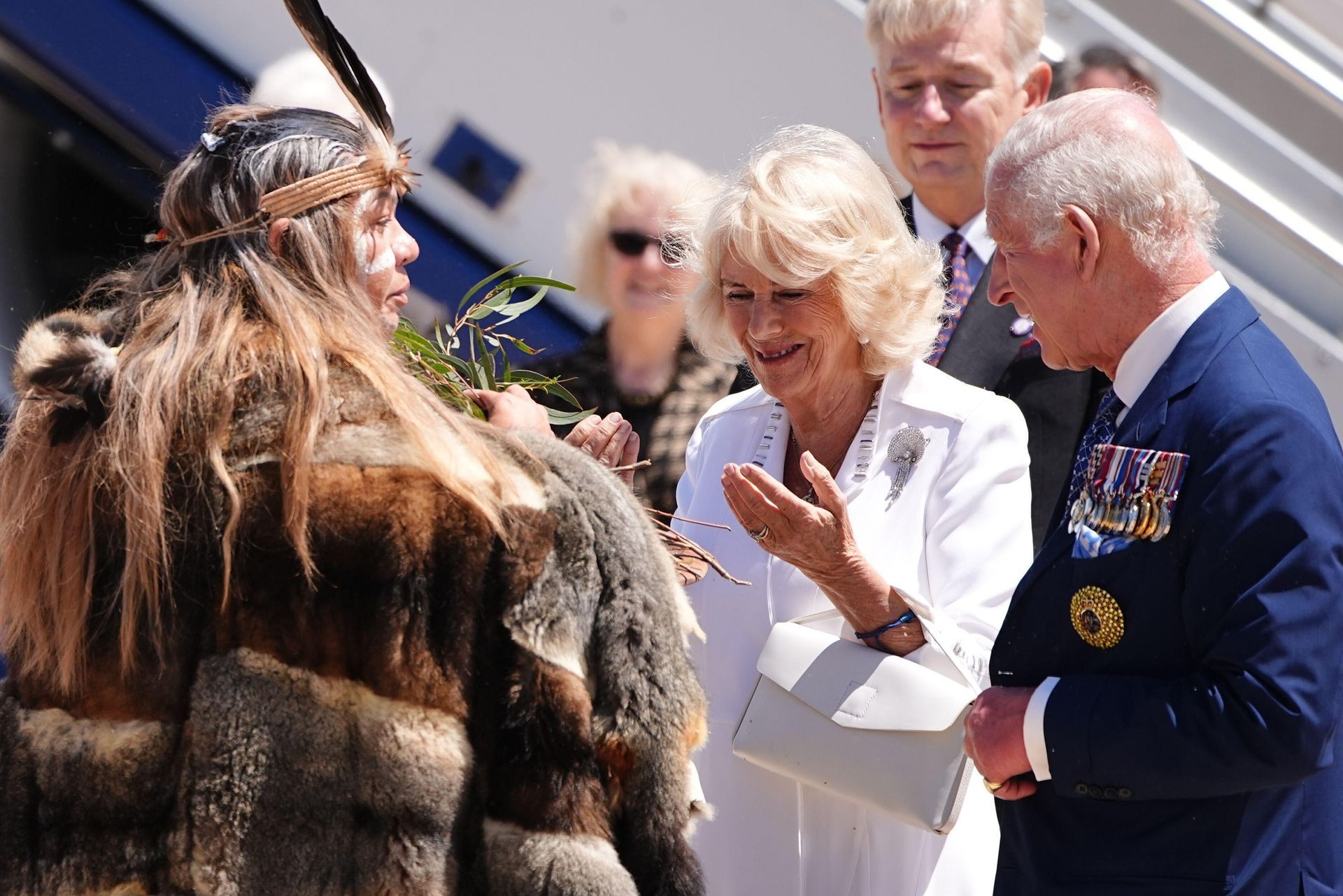 King Charles and Queen Camilla greet a person in traditional garb, sunny outdoor setting.