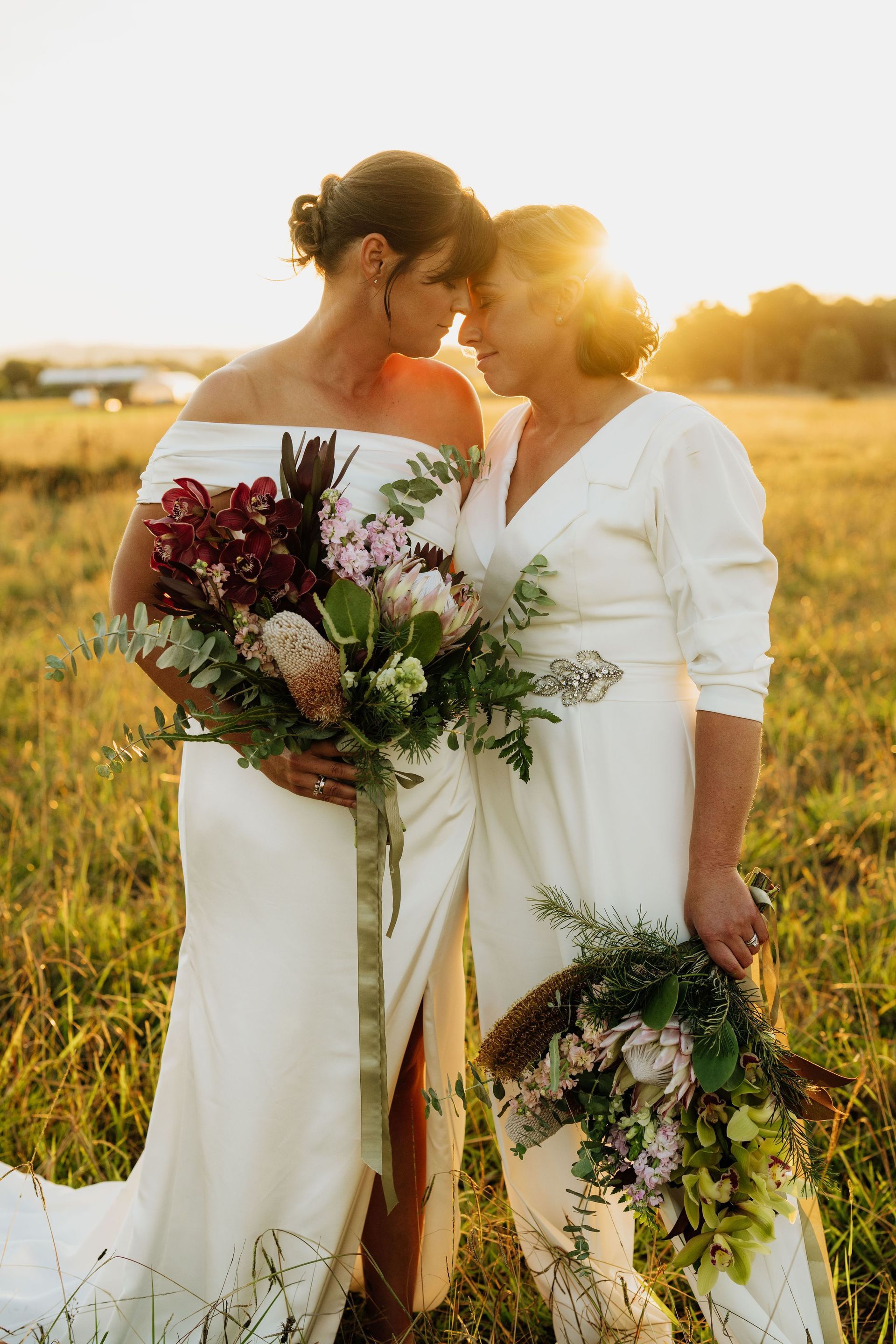 Two women in white dresses are standing next to each other in a field holding bouquets of flowers.