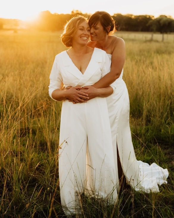 Two women in white dresses are hugging each other in a field.