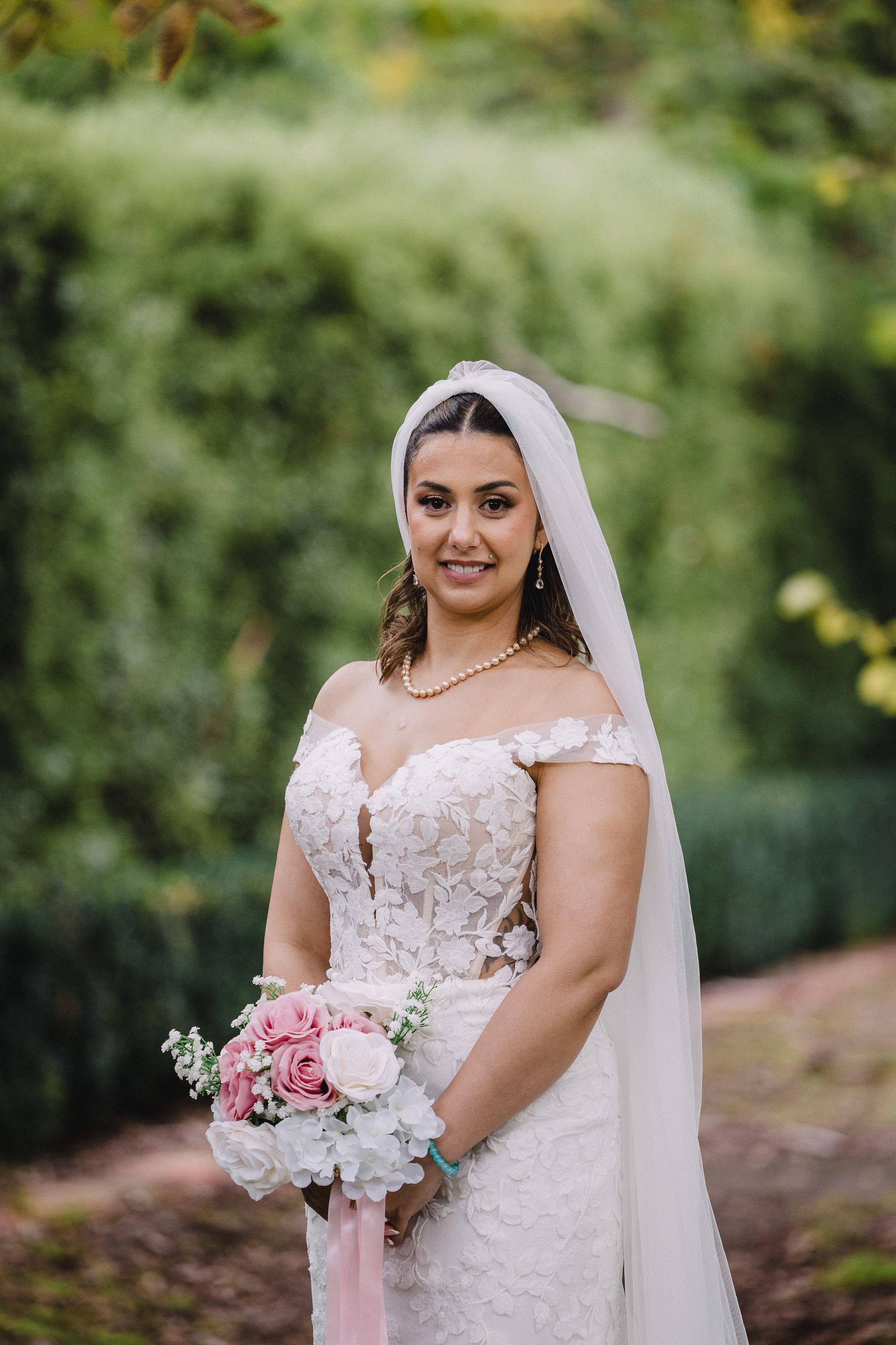 Bride in a white lace gown and veil holding a pink and white bouquet outdoors in a garden
