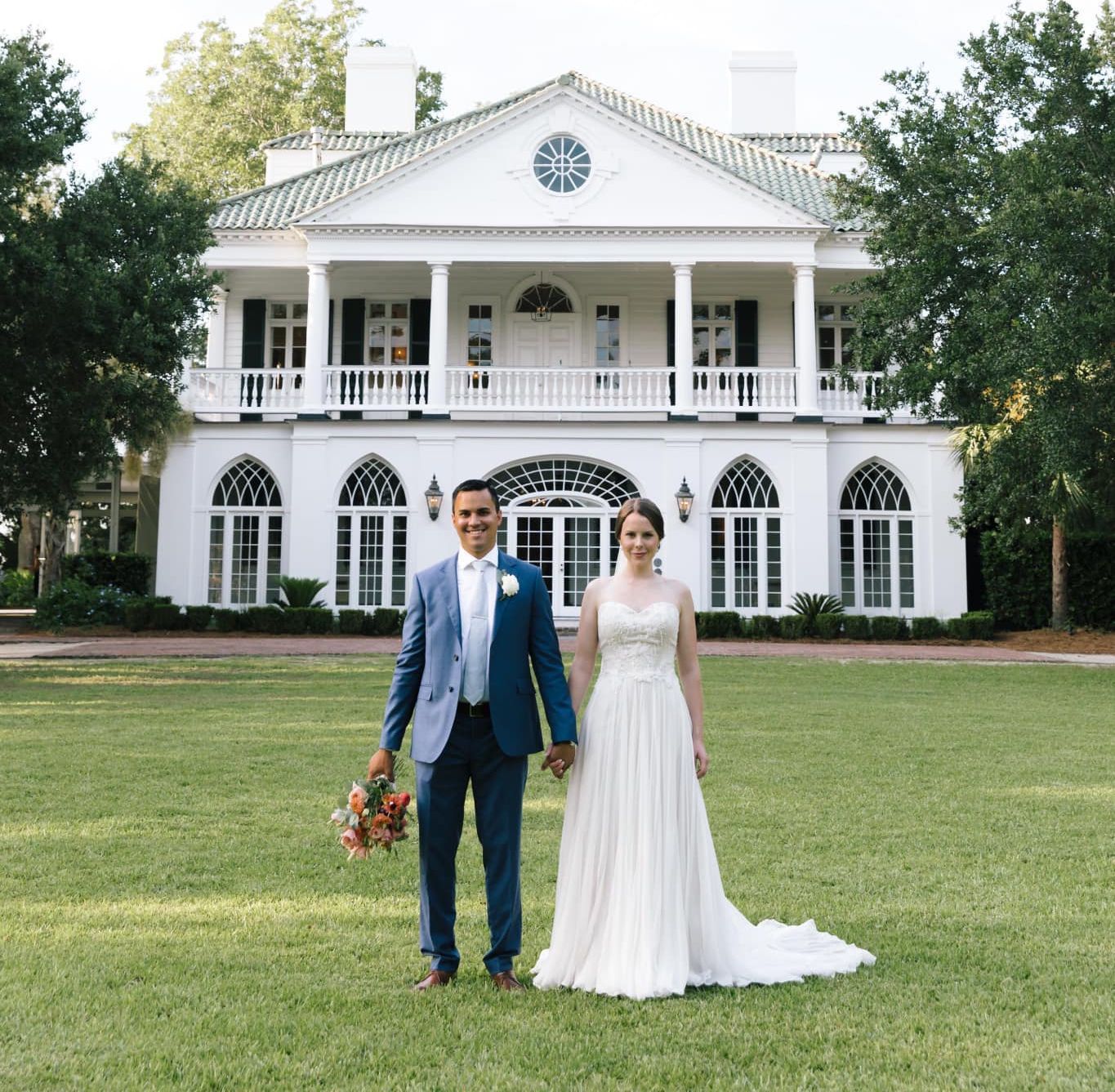 A bride and groom are standing in front of a large white house holding hands.