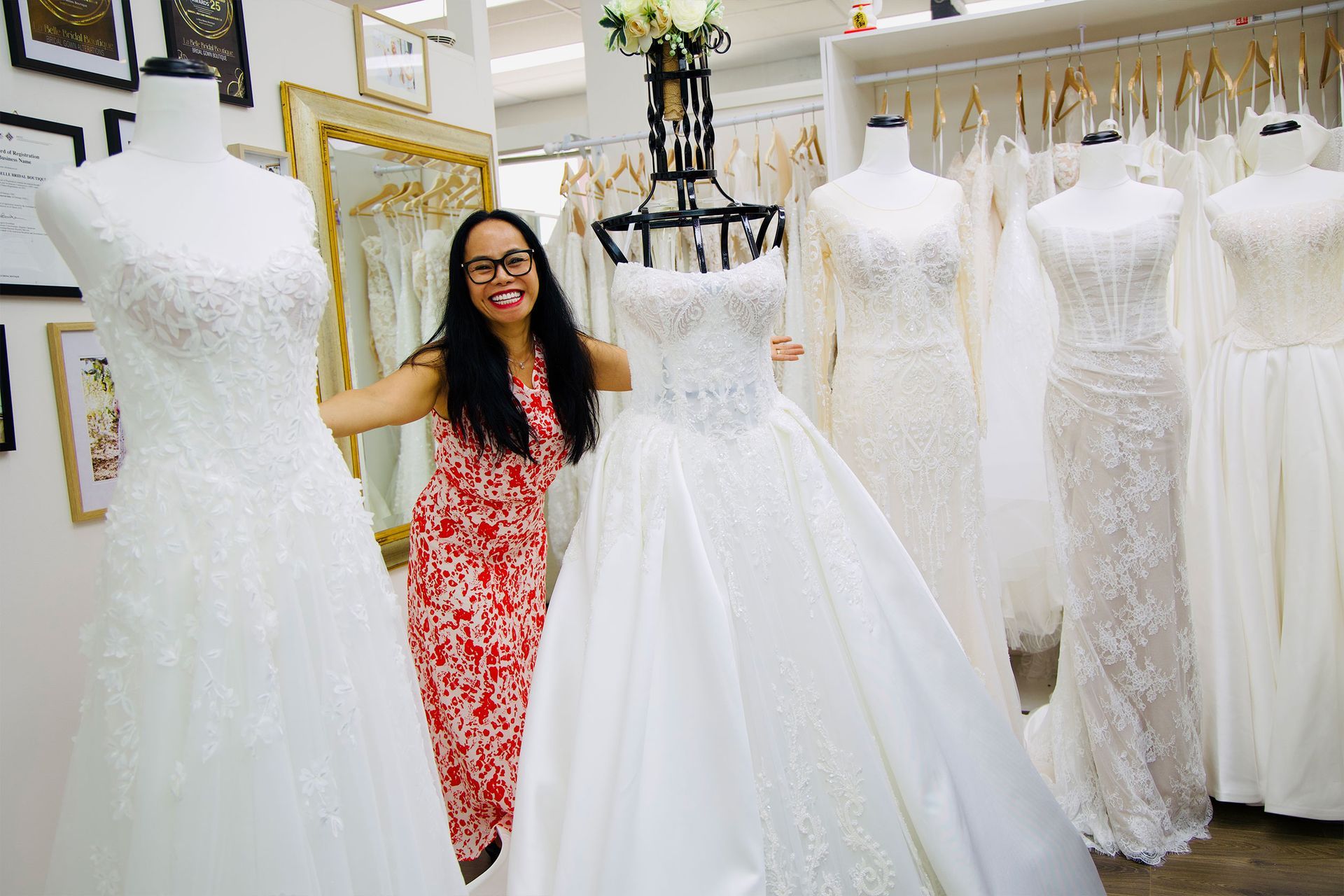 Woman smiles in a bridal shop, posing by a wedding dress on a mannequin; other dresses in the background.
