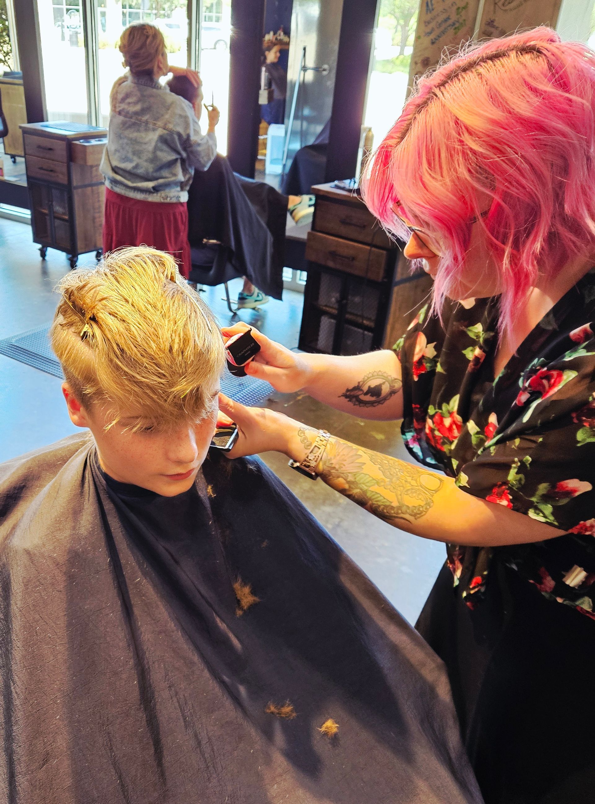 A person with pink hair is cutting a young person's hair in a salon. Both have speckled hair.