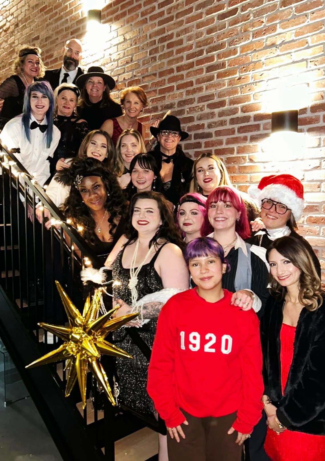 Group photo on staircase. People in festive attire. Brick wall backdrop. Golden star decor.