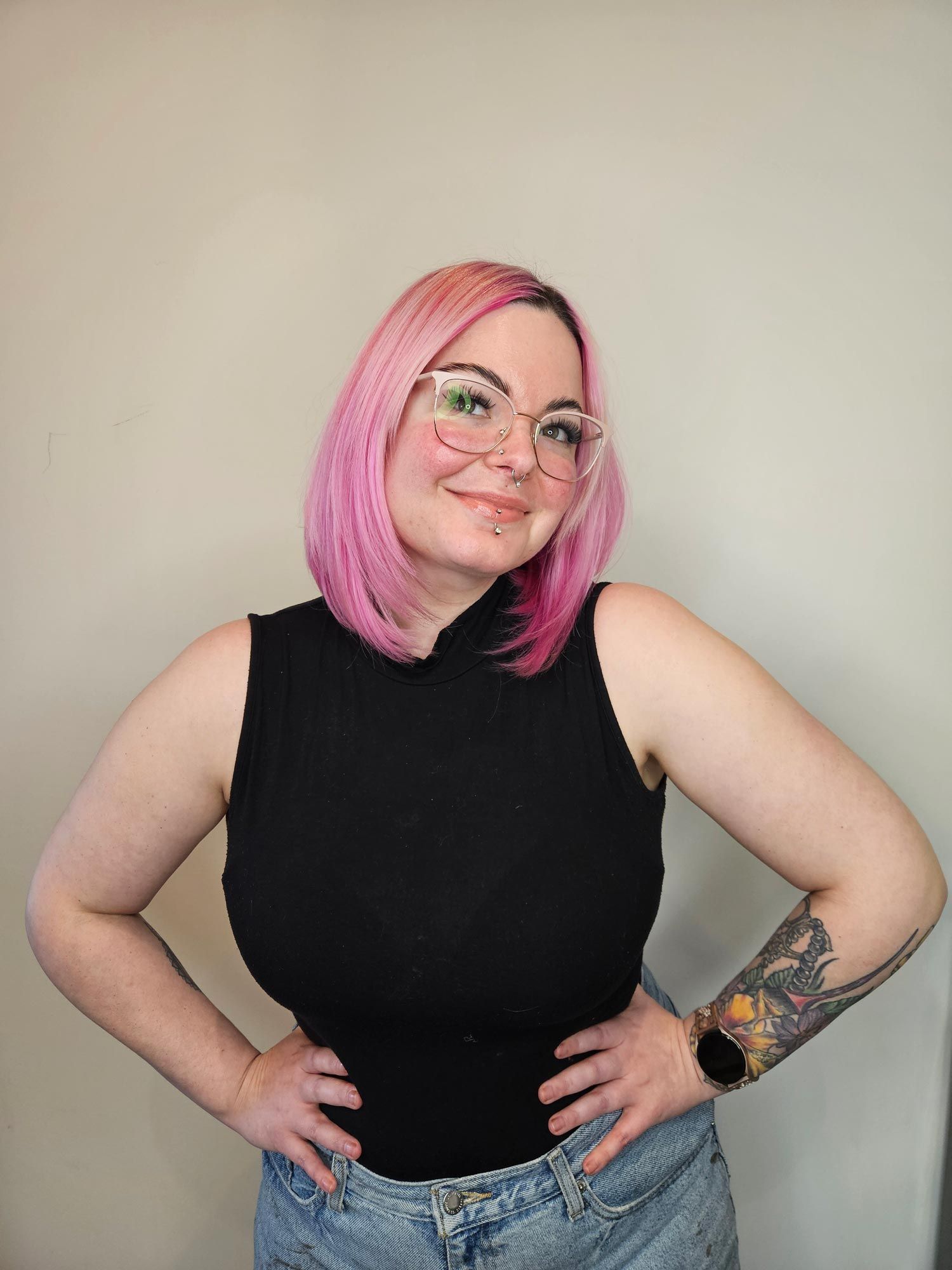 Woman with pink hair and glasses, wearing a black sleeveless top and jean shorts, hands on hips, smiling, tattoo on arm.