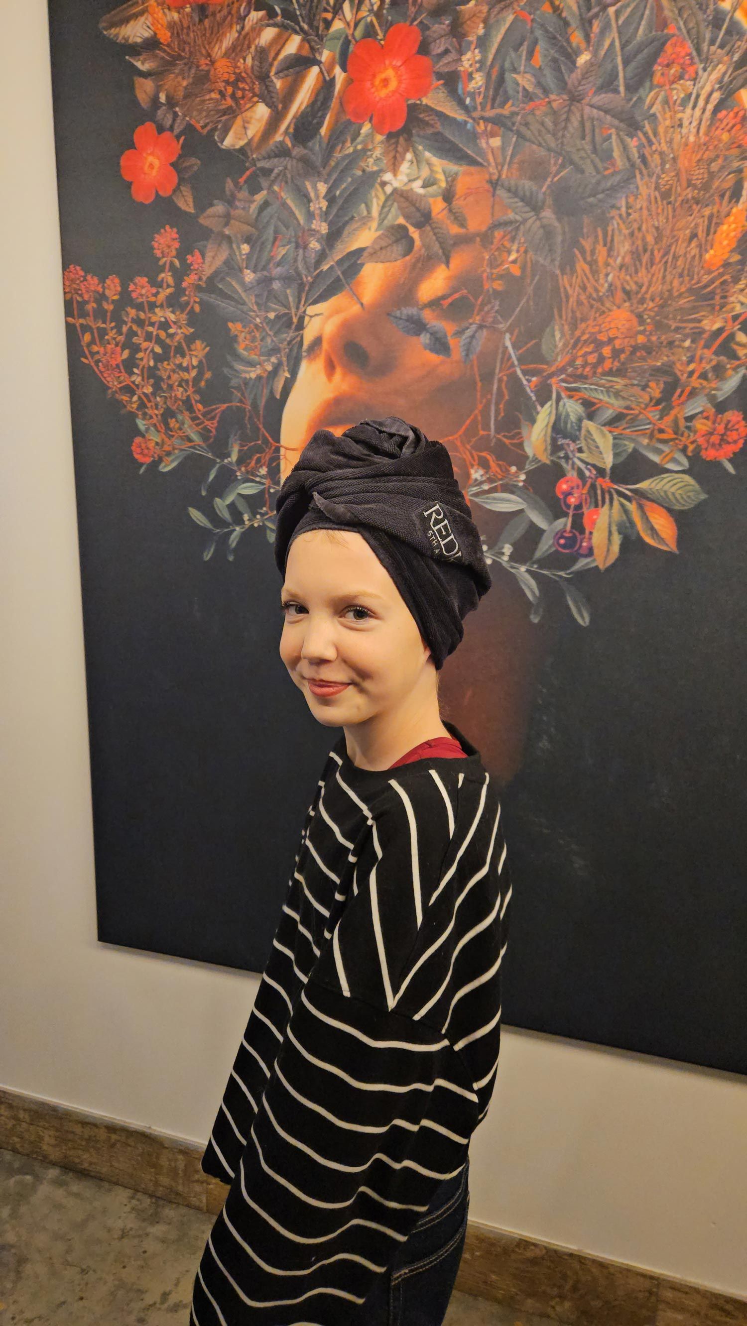 Girl wearing a black turban and striped shirt smiles in front of floral artwork.