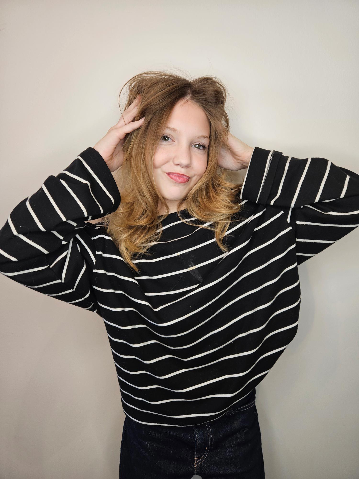 Person in striped black and white sweater, hands in hair, smiling.