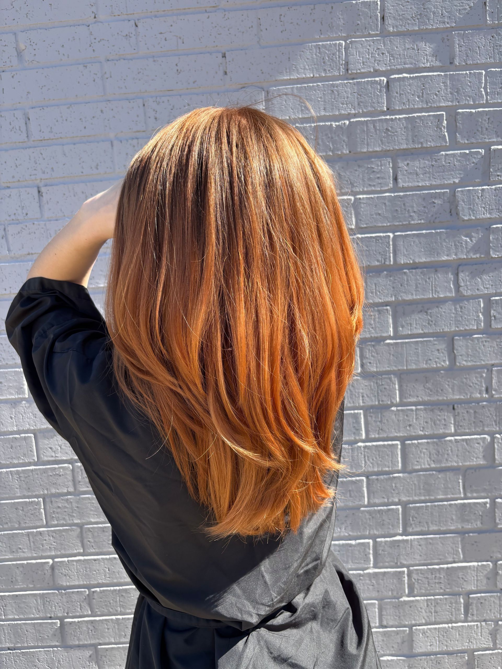 Person with long, reddish-brown hair stands in front of a white brick wall. Back view, hands near head.
