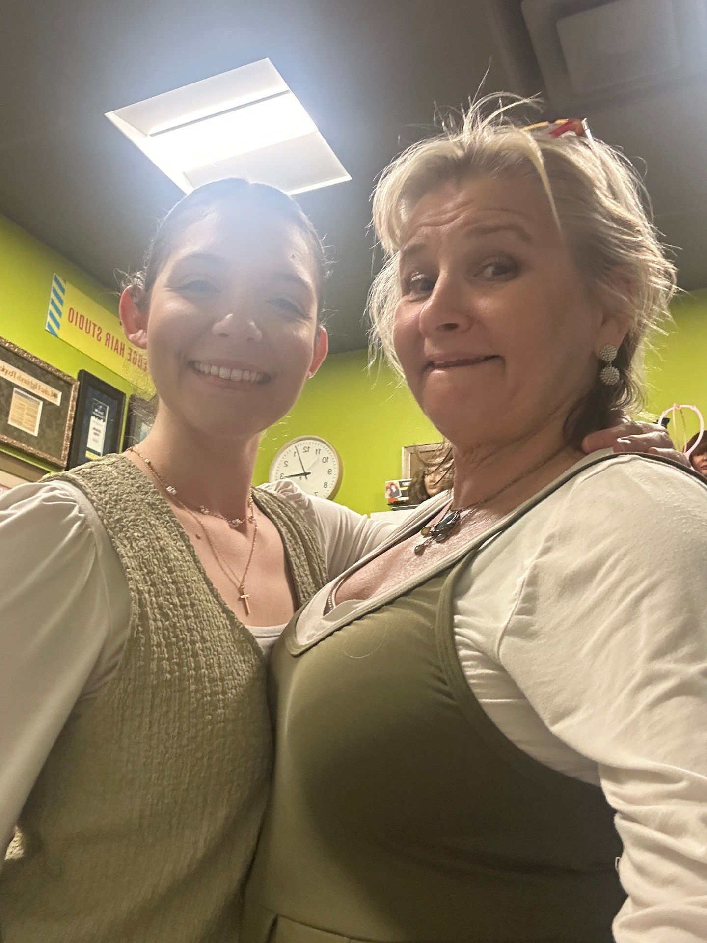 Two women smiling, posing for a selfie indoors. One has light hair, the other dark. Green wall in background.