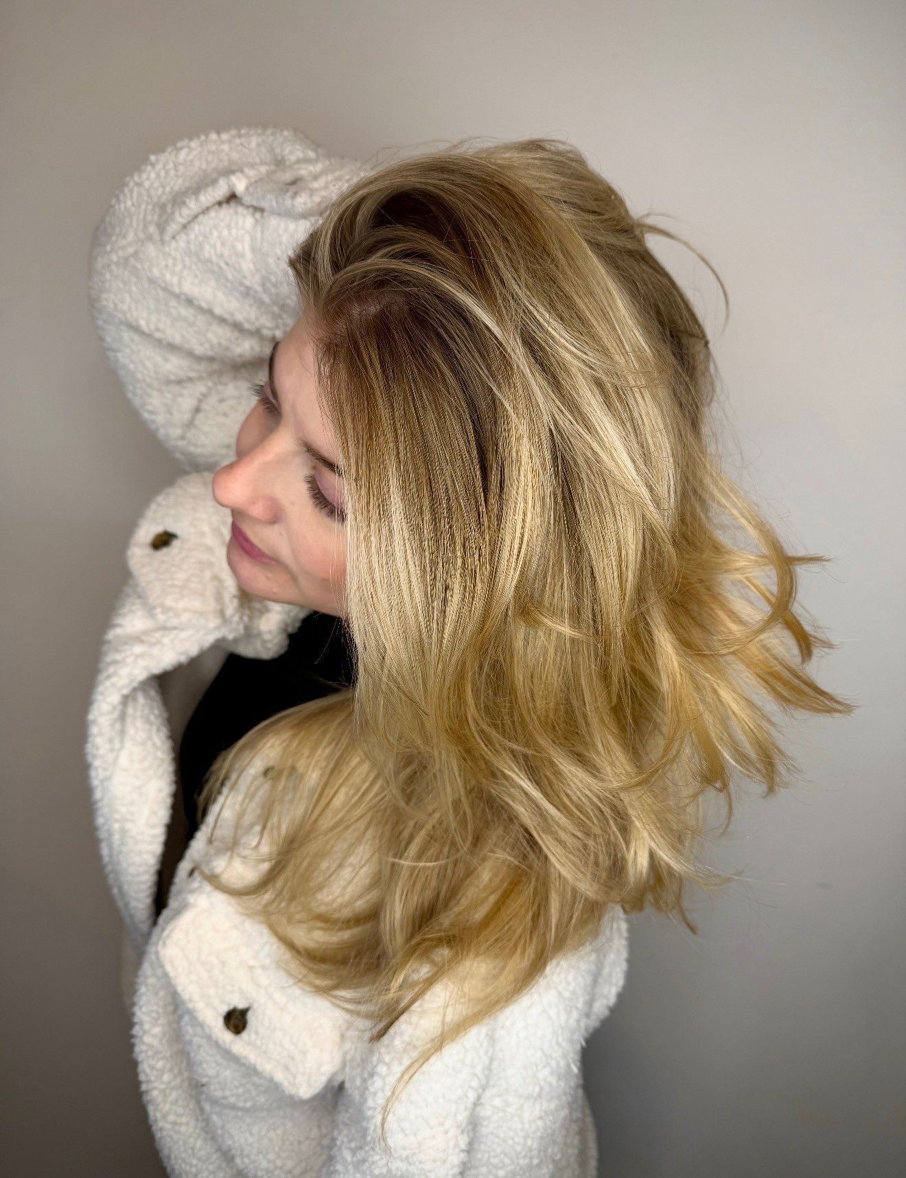 Woman in a fluffy white jacket with blonde hair, looking upwards, near a plain white wall.