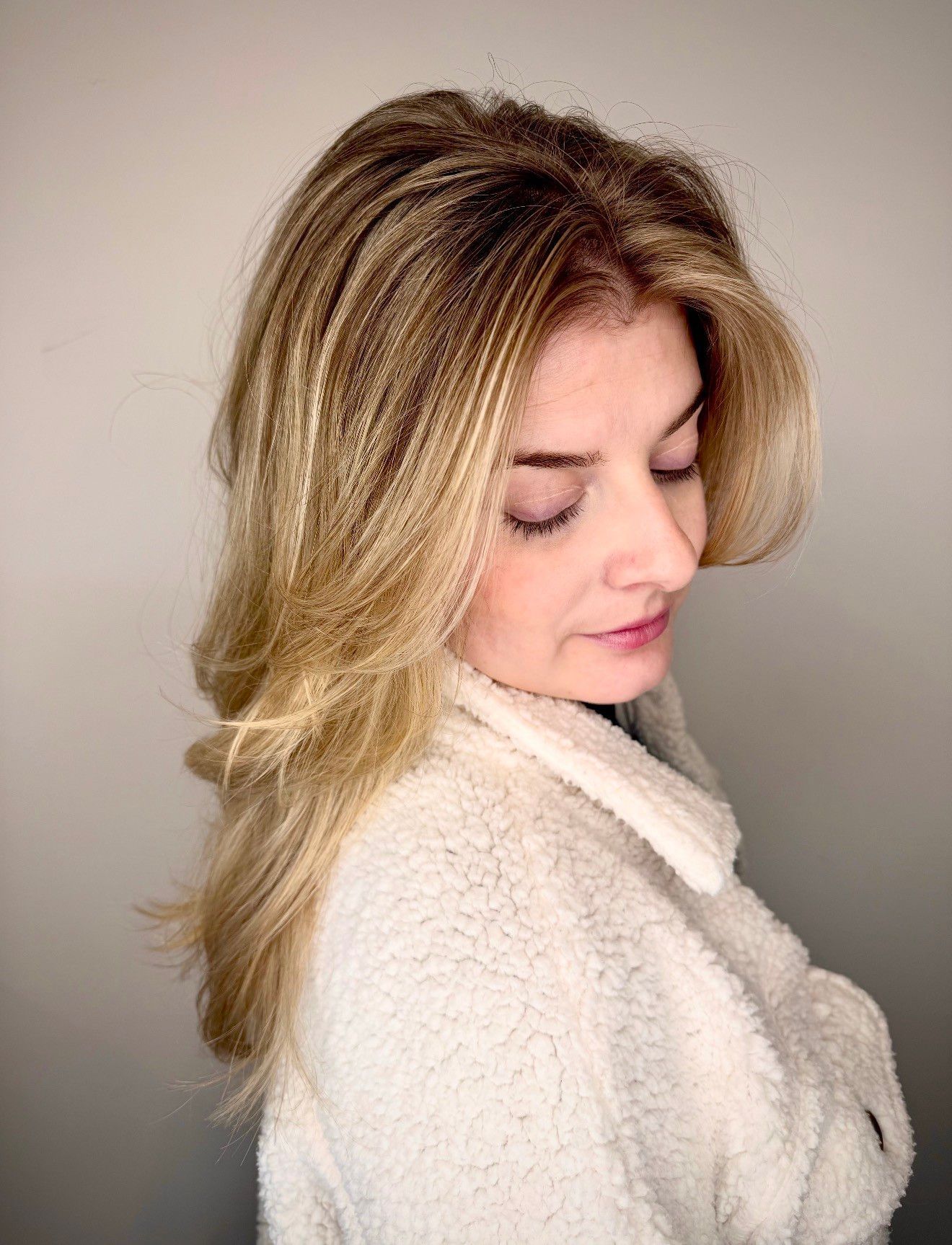 Woman with layered blonde hair, wearing a white fluffy jacket, looking down.