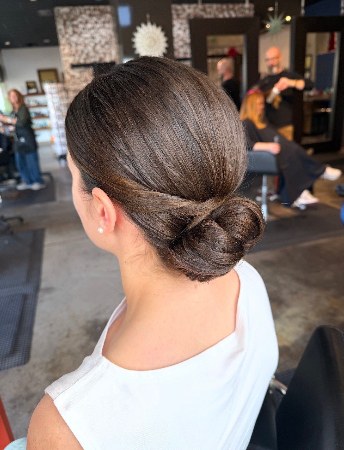Woman with dark brown hair in a low bun at a salon, wearing a white shirt.
