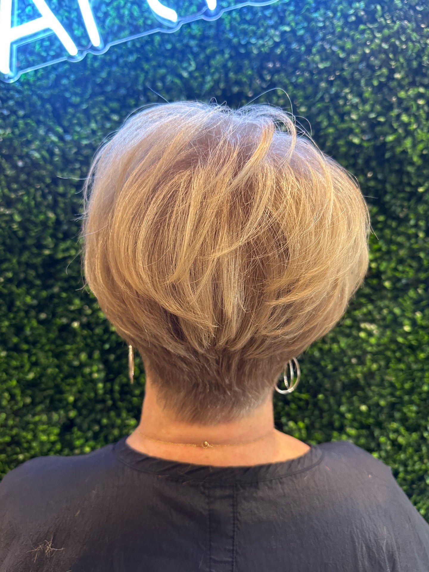 Back view of a person with a short, layered blonde haircut against a green hedge wall.