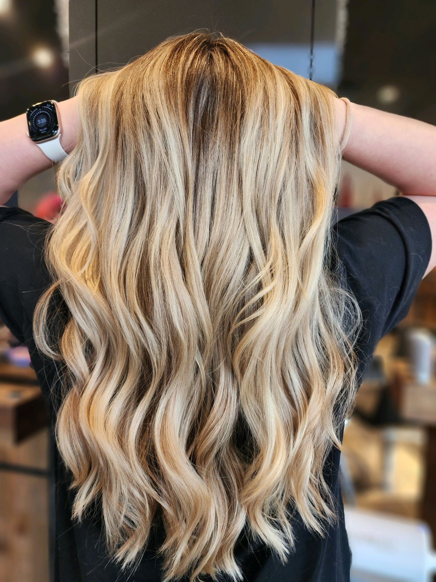 Blonde hair with balayage, styled in waves. Hands raised, wrists visible with watch.