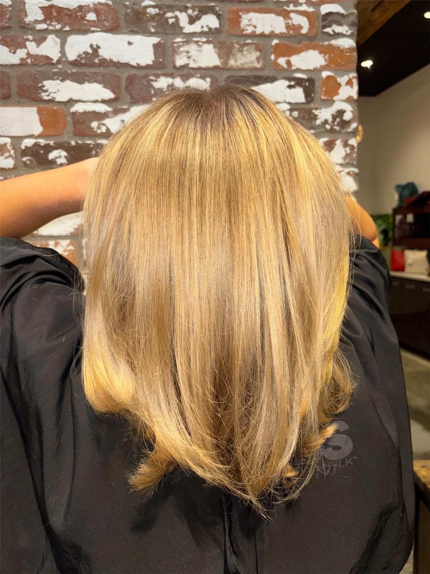 Woman with layered blonde hair at a salon. The hair is light, the background is a brick wall.